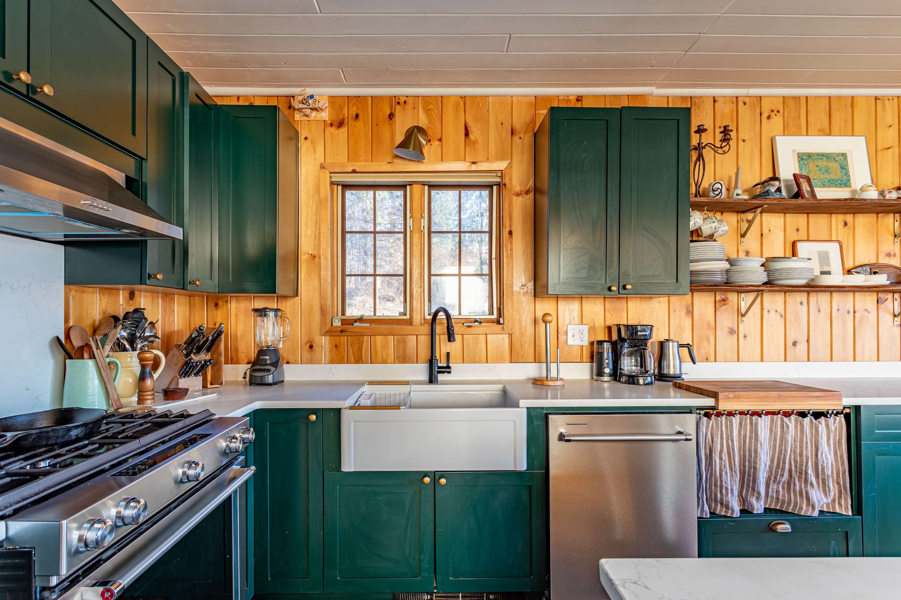 Detailed view of the kitchen counter area with a white farmhouse sink, black faucet, stainless steel dishwasher, and dark green lower cabinets against natural wood paneled walls.
