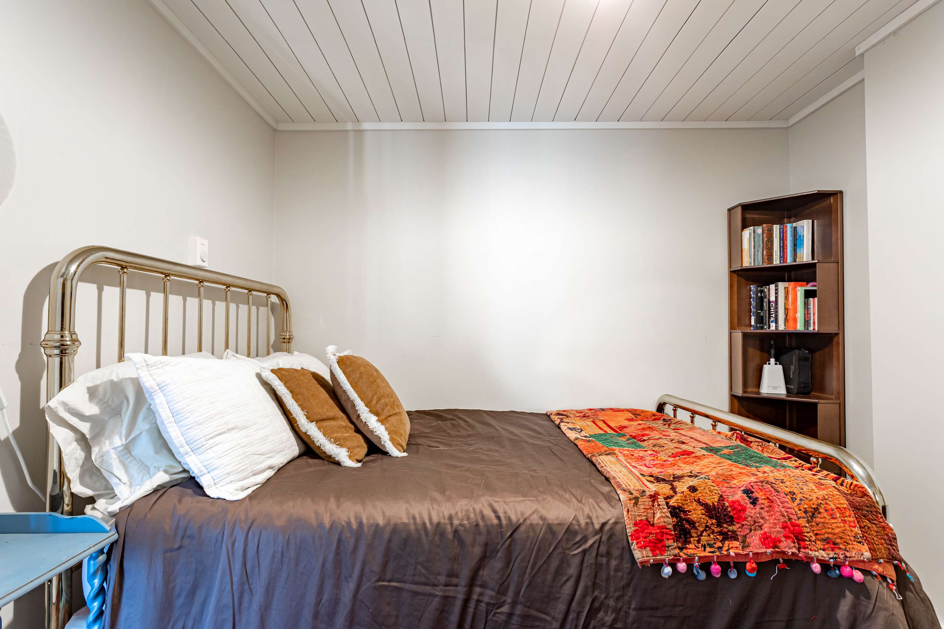 A cozy bedroom with light gray walls and a white paneled ceiling, showcasing a metal bed frame with brown bedding and white and brown pillows, and a corner bookshelf.