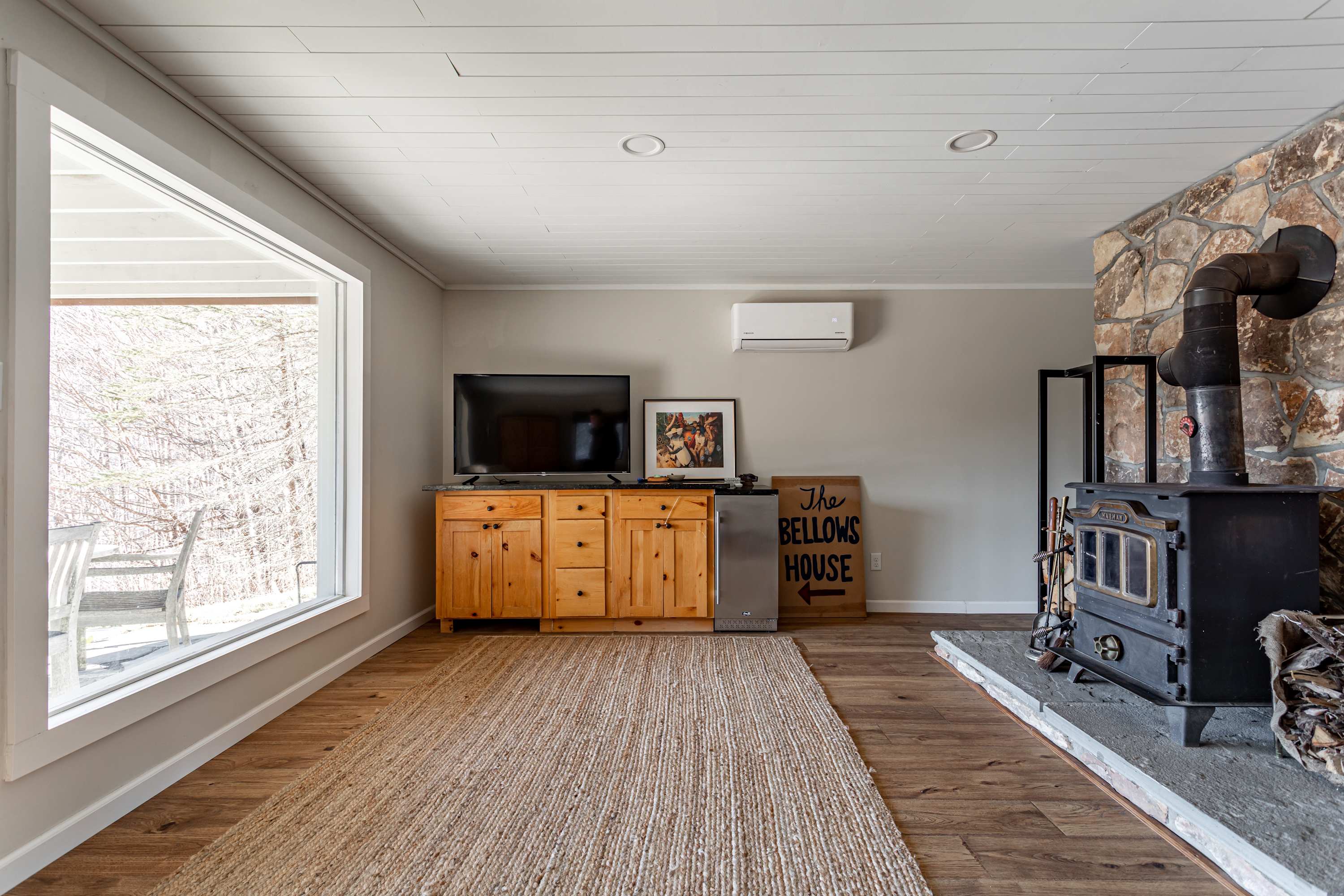 Cozy living room featuring a wood-burning stove with a stone hearth, a wooden TV console, and a large window offering views of the surrounding trees. A jute rug covers the light wood floor, with a mini-split AC unit mounted above.