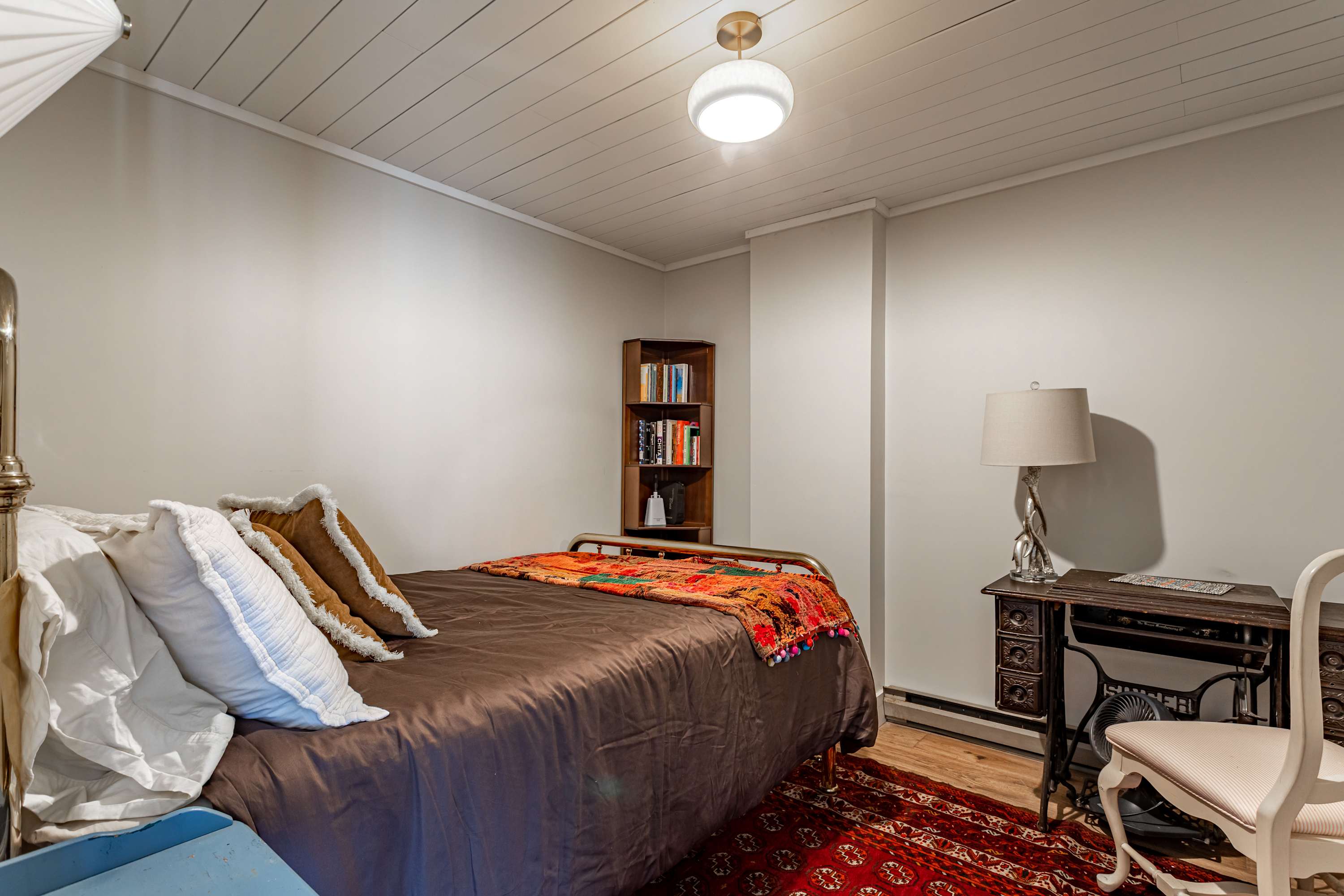 A simple and bright bedroom with light gray walls and a white paneled ceiling, featuring a metal bed frame with brown bedding and colorful throw pillows, and a corner bookshelf.