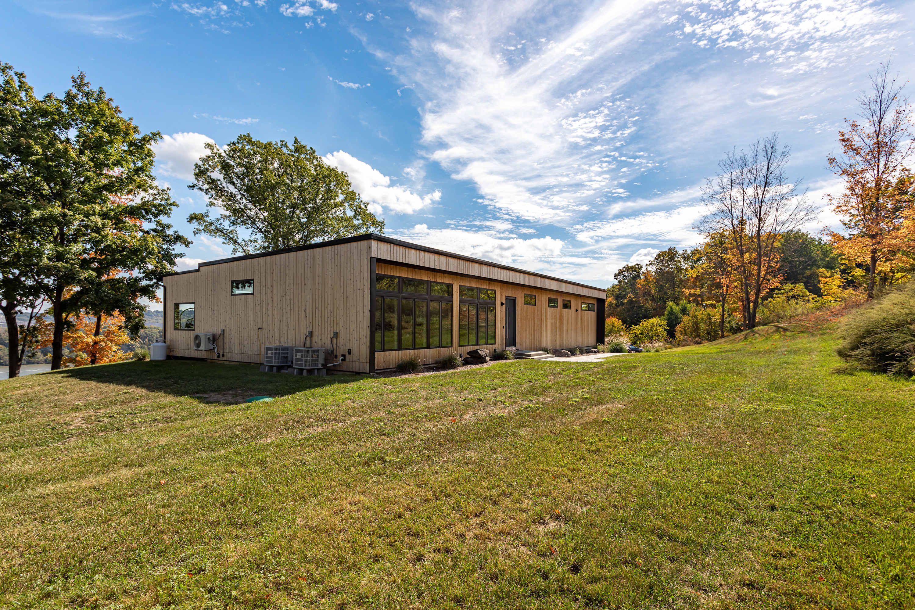 Side exterior view of the modern house, showcasing its light wood siding, large windows, and a flat roof, situated on a grassy slope with trees and a clear blue sky overhead.