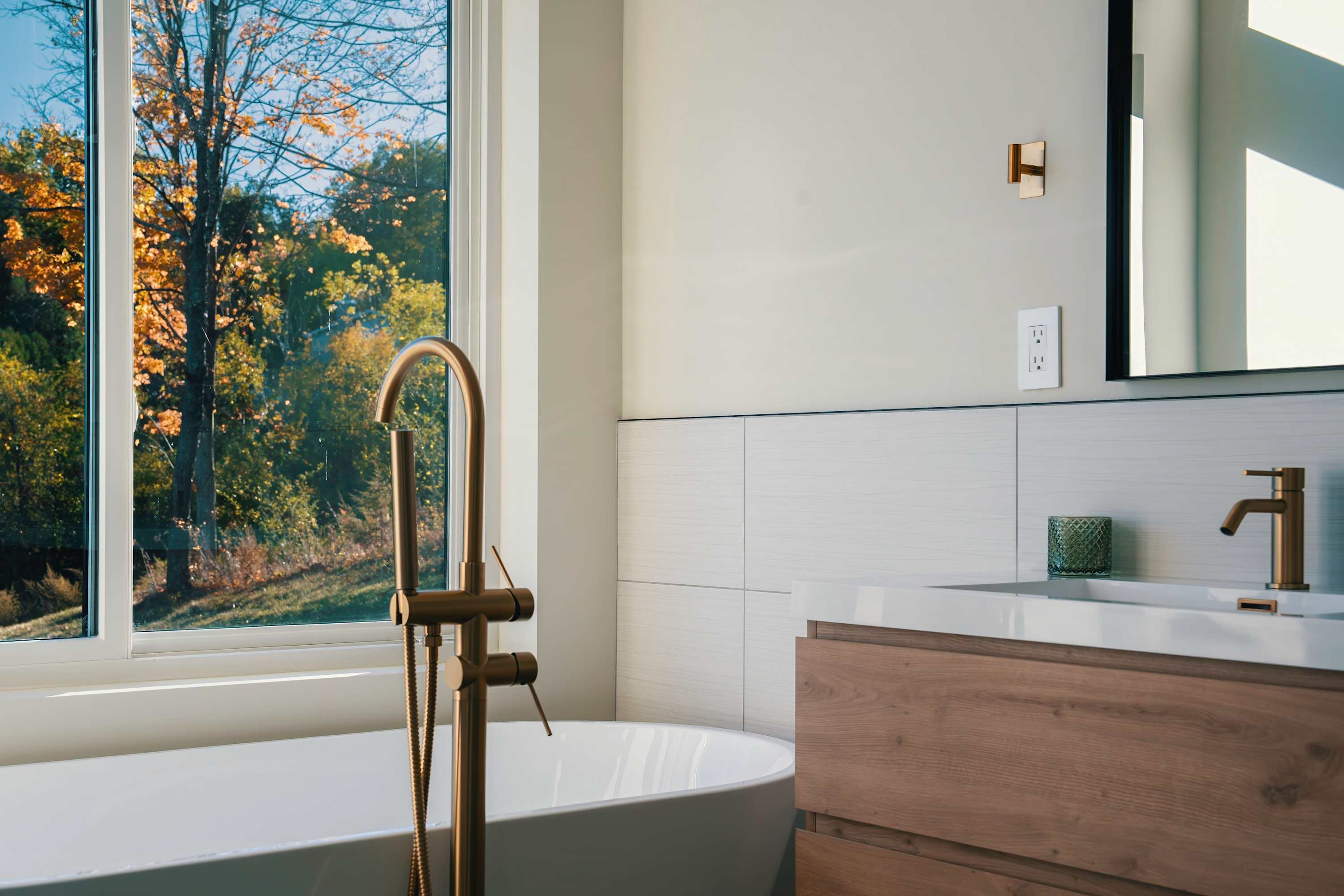 Modern bathroom featuring a freestanding white bathtub with a brass floor-mounted faucet, a wooden vanity with a white countertop, and a large window offering views of colorful autumn trees.