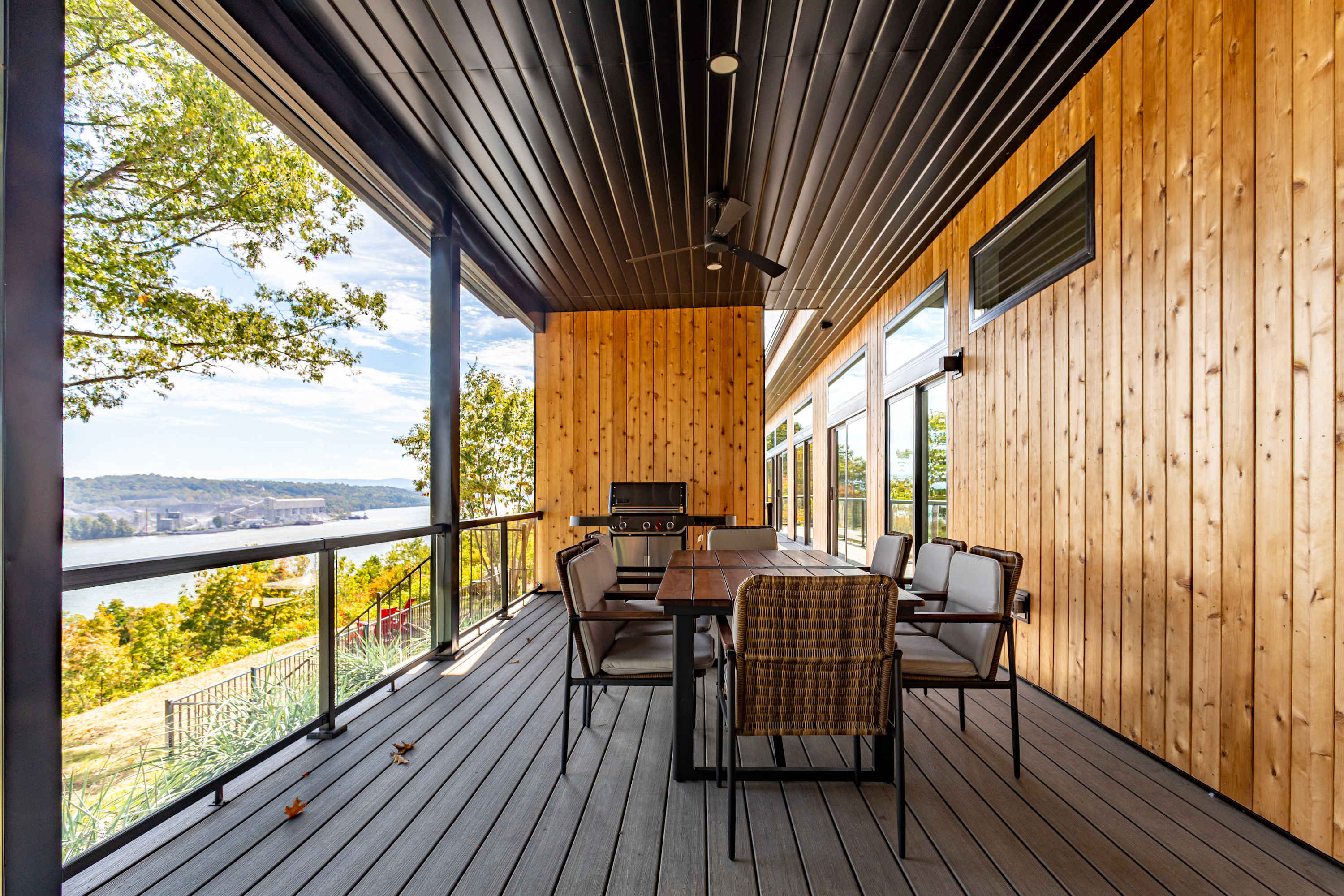 Expansive outdoor deck with a dark slatted ceiling and floor, featuring a dining table with chairs, a BBQ grill, and glass railings offering panoramic views of a wide river and distant hills.