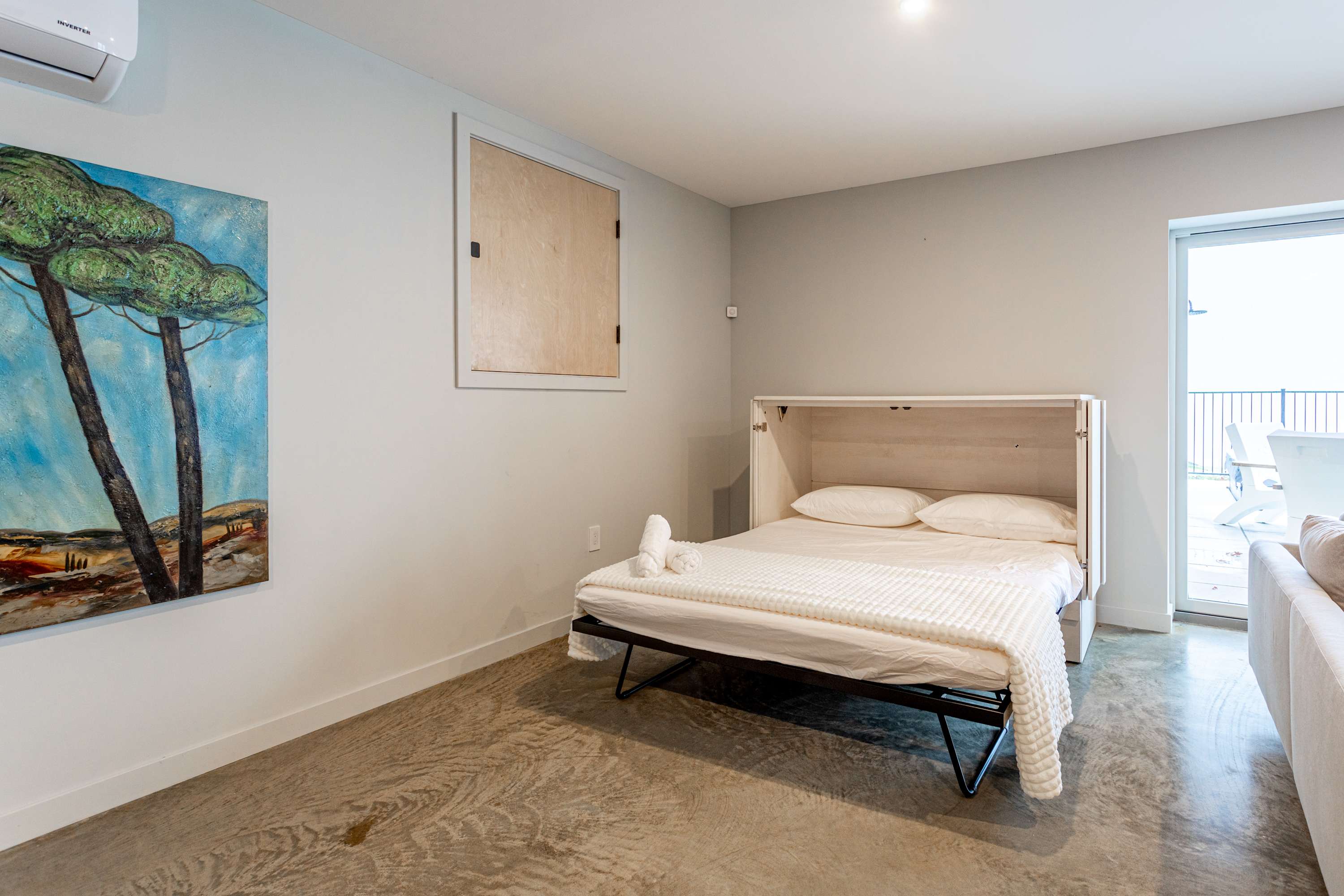 A bright room features a white fold-out bed with white bedding and towels, set against light gray walls and a concrete floor. A large abstract painting with green trees and a blue sky hangs on the left, with an AC unit above it.