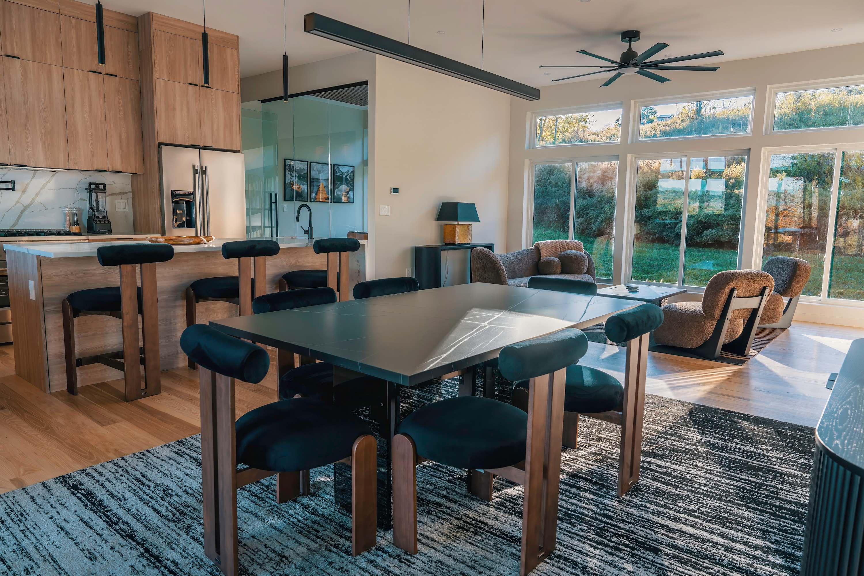 Open-concept dining area with a sleek black dining table and velvet chairs, adjacent to a modern kitchen with light wood cabinetry and a breakfast bar, all illuminated by natural light from large windows.