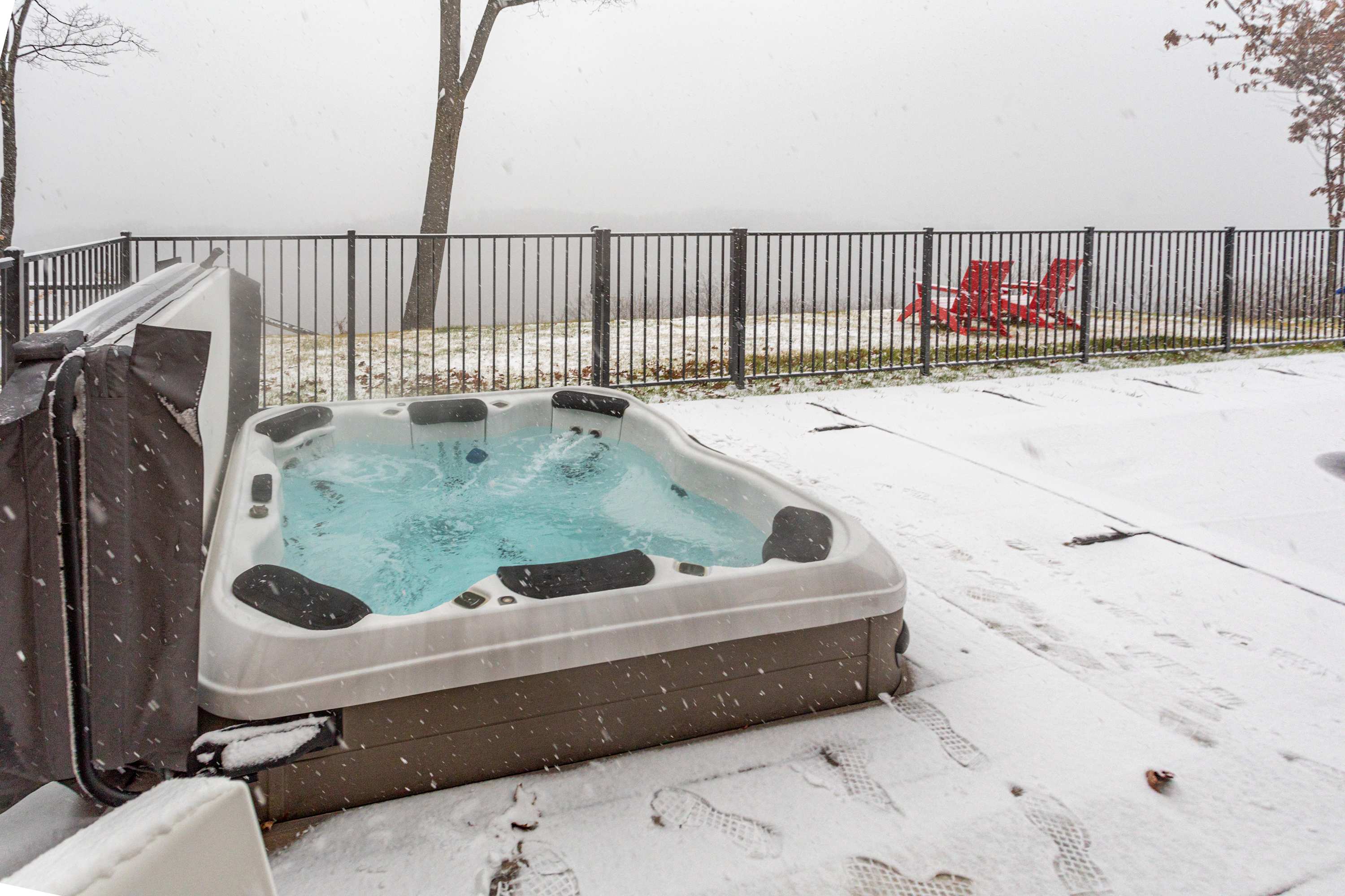 A hot tub with bubbling blue water sits on a snow-dusted patio, with a black metal fence separating it from a distant, misty landscape. Two red Adirondack chairs are visible beyond the fence, adding a splash of color to the winter setting.