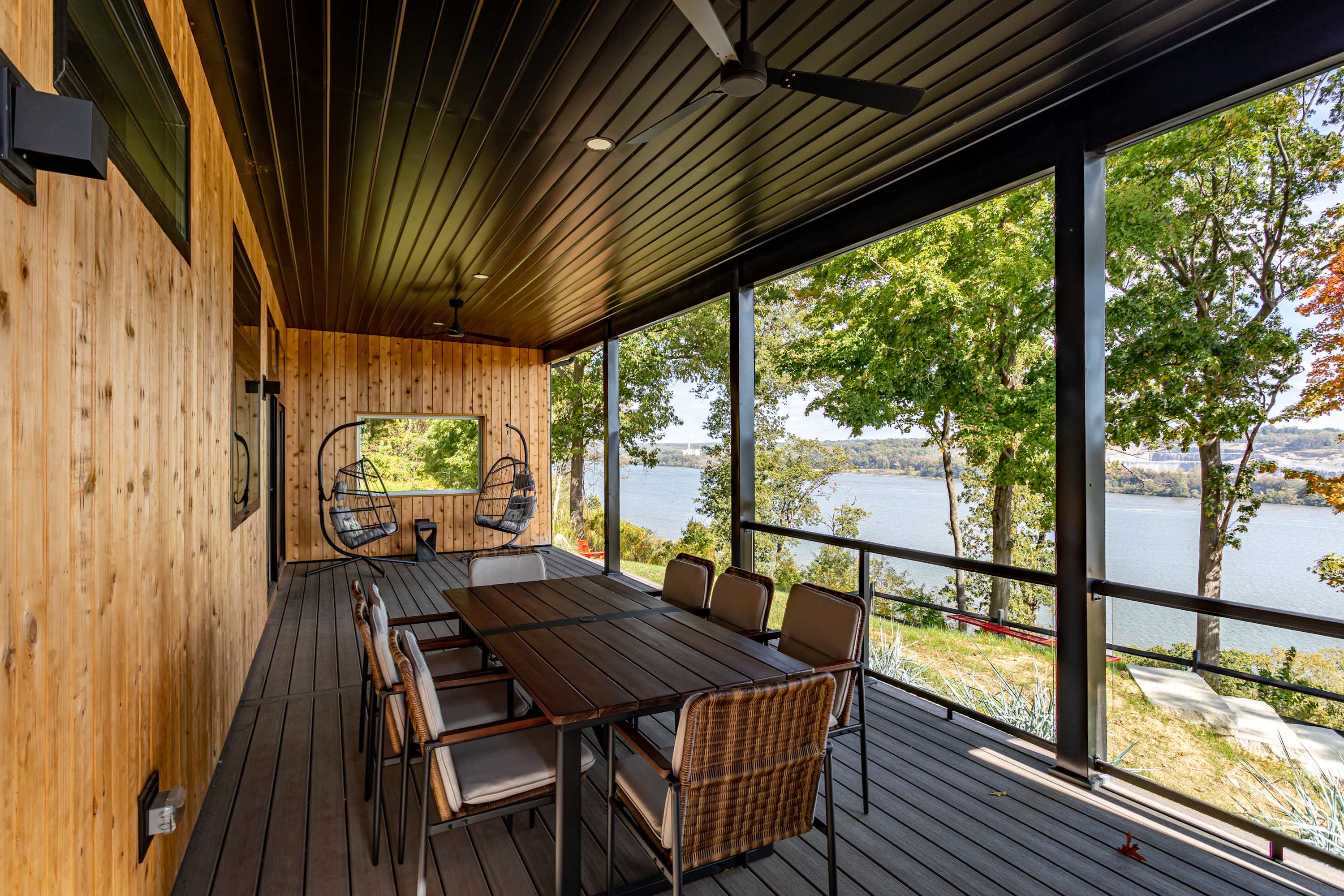 Spacious covered deck with a long wooden dining table and chairs, offering panoramic river and tree views. The ceiling is dark wood, and the walls are light wood, with two hanging egg chairs in the background.