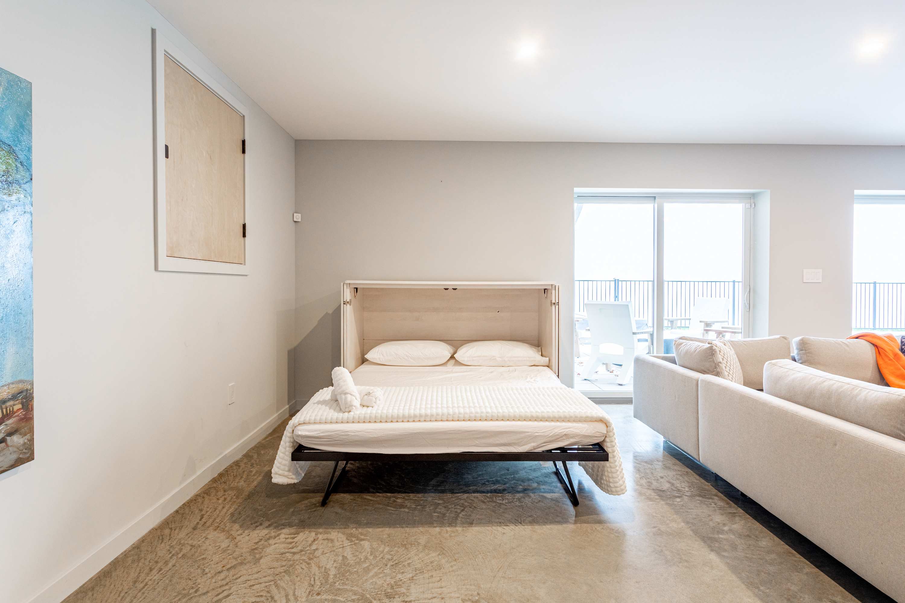A functional secondary living space with a murphy bed pulled down and made up, offering an additional sleeping area, adjacent to a sliding glass door that opens to an outdoor balcony, all on a concrete floor.