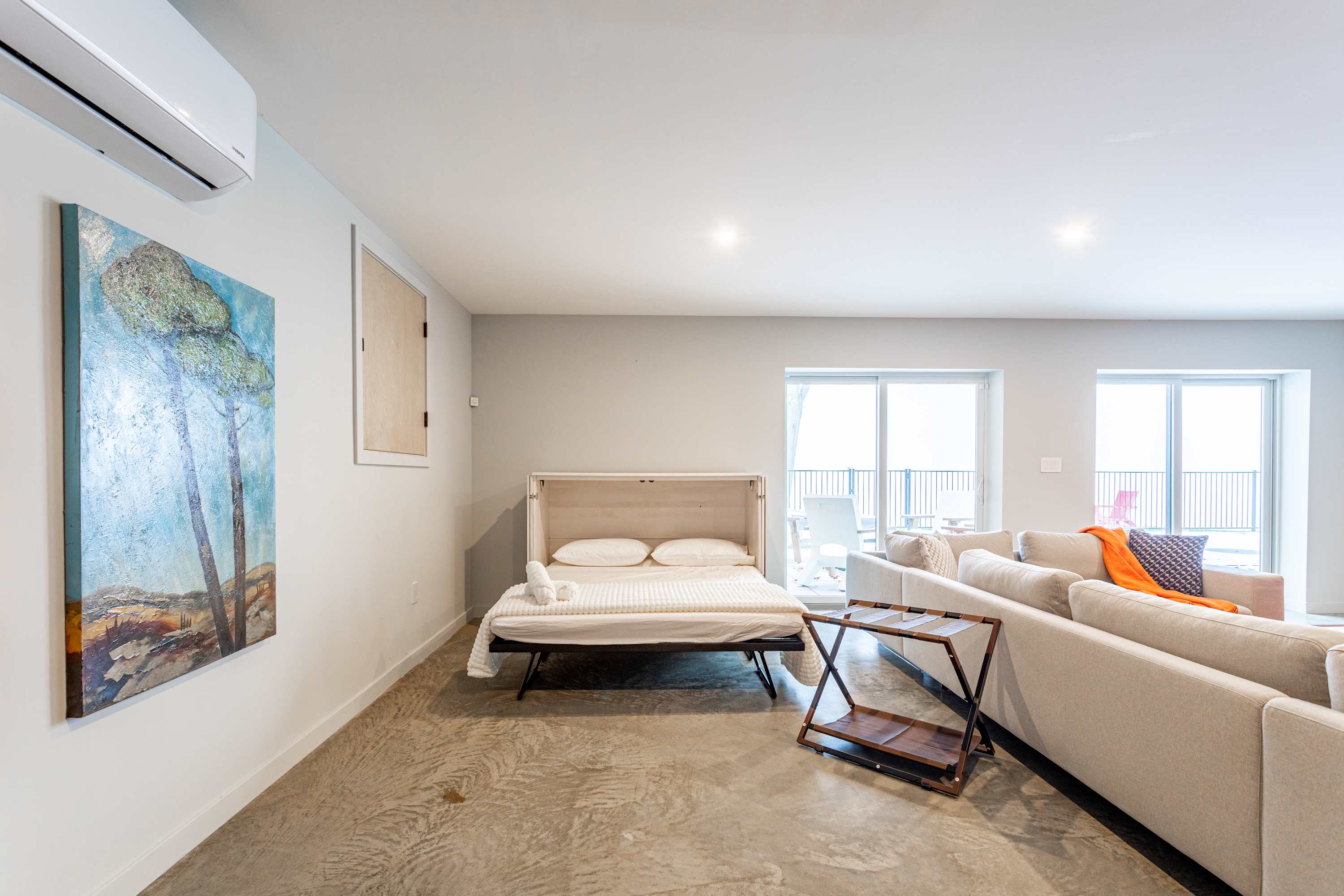 A spacious living area with light gray walls and a concrete floor, featuring a white fold-out bed with white bedding and a light-colored sectional sofa. A large sliding glass door on the right opens to an outdoor space, while an abstract painting and AC unit are visible on the left.