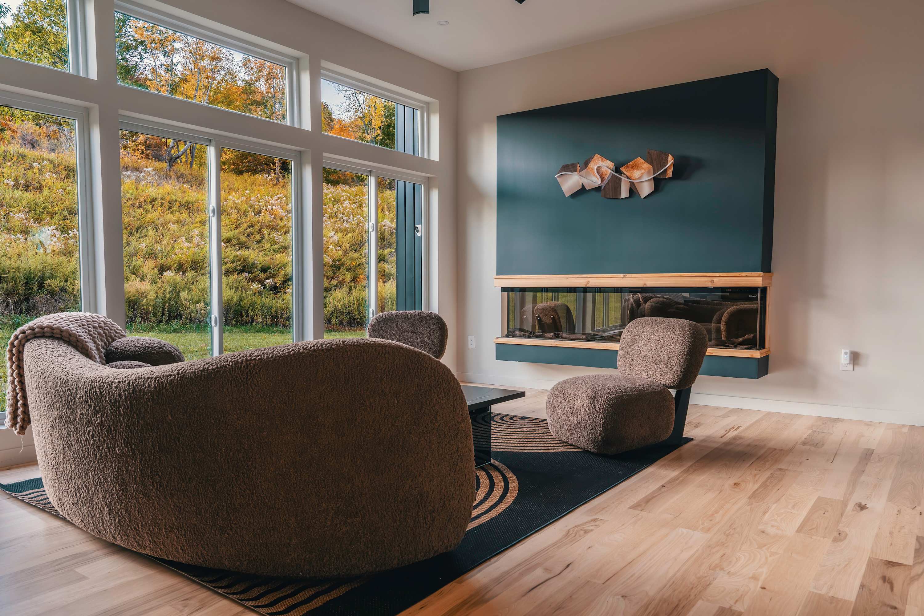 A bright living room with a curved brown sofa and matching armchairs arranged on a patterned rug, facing a large window wall that offers expansive views of an autumn landscape, with a dark teal fireplace unit on the right.