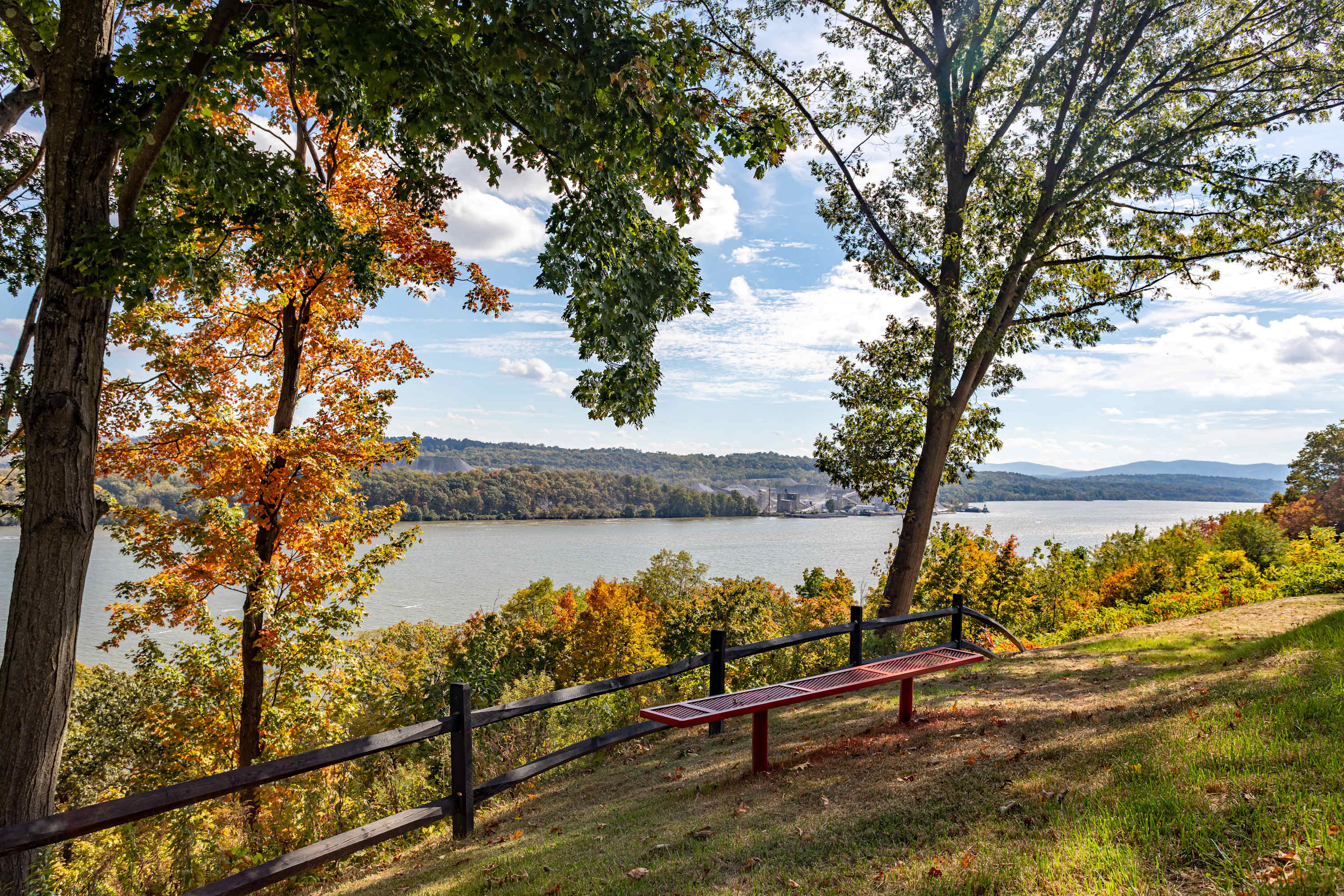 Picturesque outdoor scene showcasing a vibrant autumn landscape with trees displaying fall colors, a red bench, and a black fence, all overlooking a wide river under a blue sky with scattered clouds.