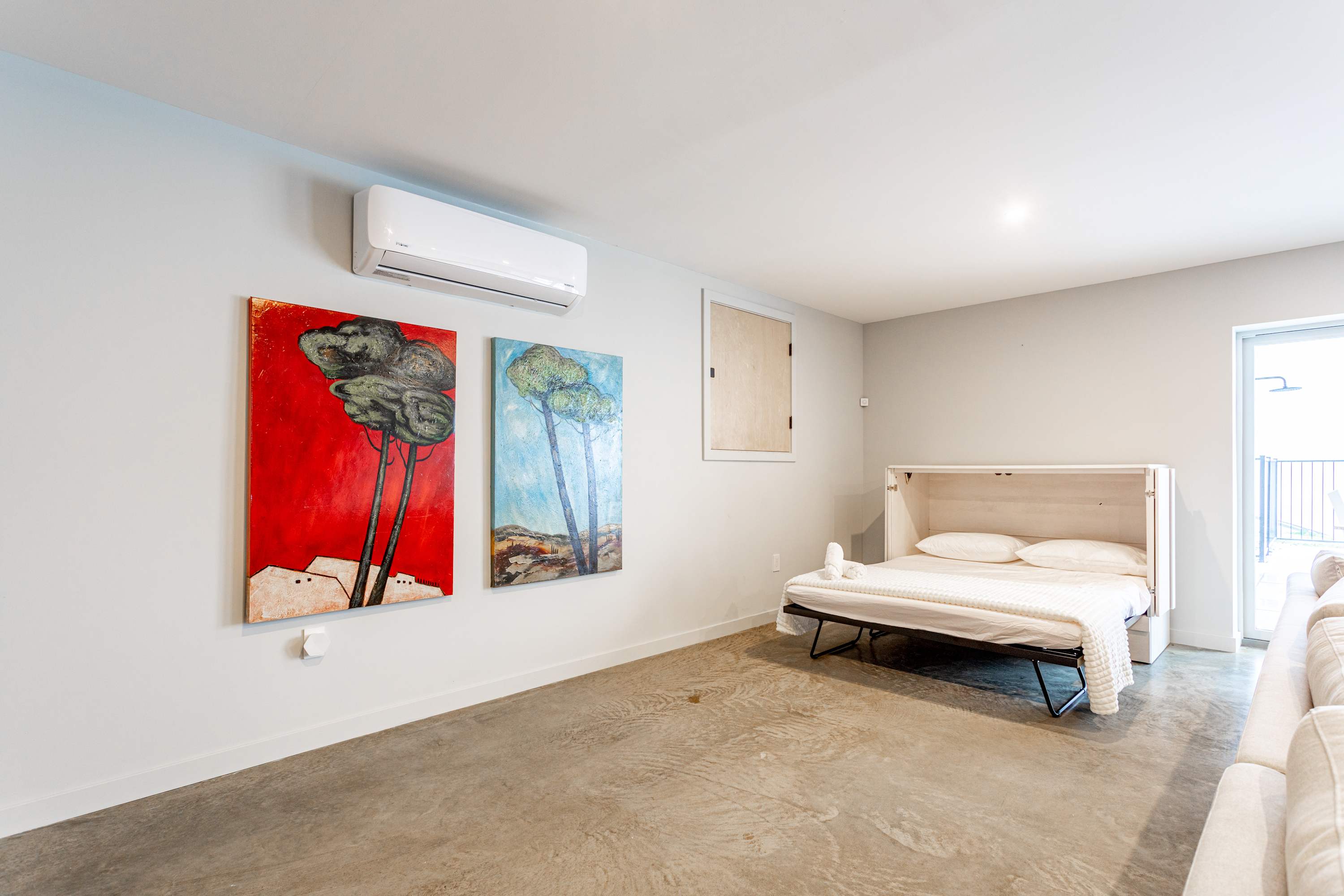 A modern living space with light gray walls and a concrete floor, featuring a white fold-out bed with white bedding. Two vibrant abstract paintings, one red and one blue, hang on the left wall below an AC unit, adding a pop of color to the minimalist design.