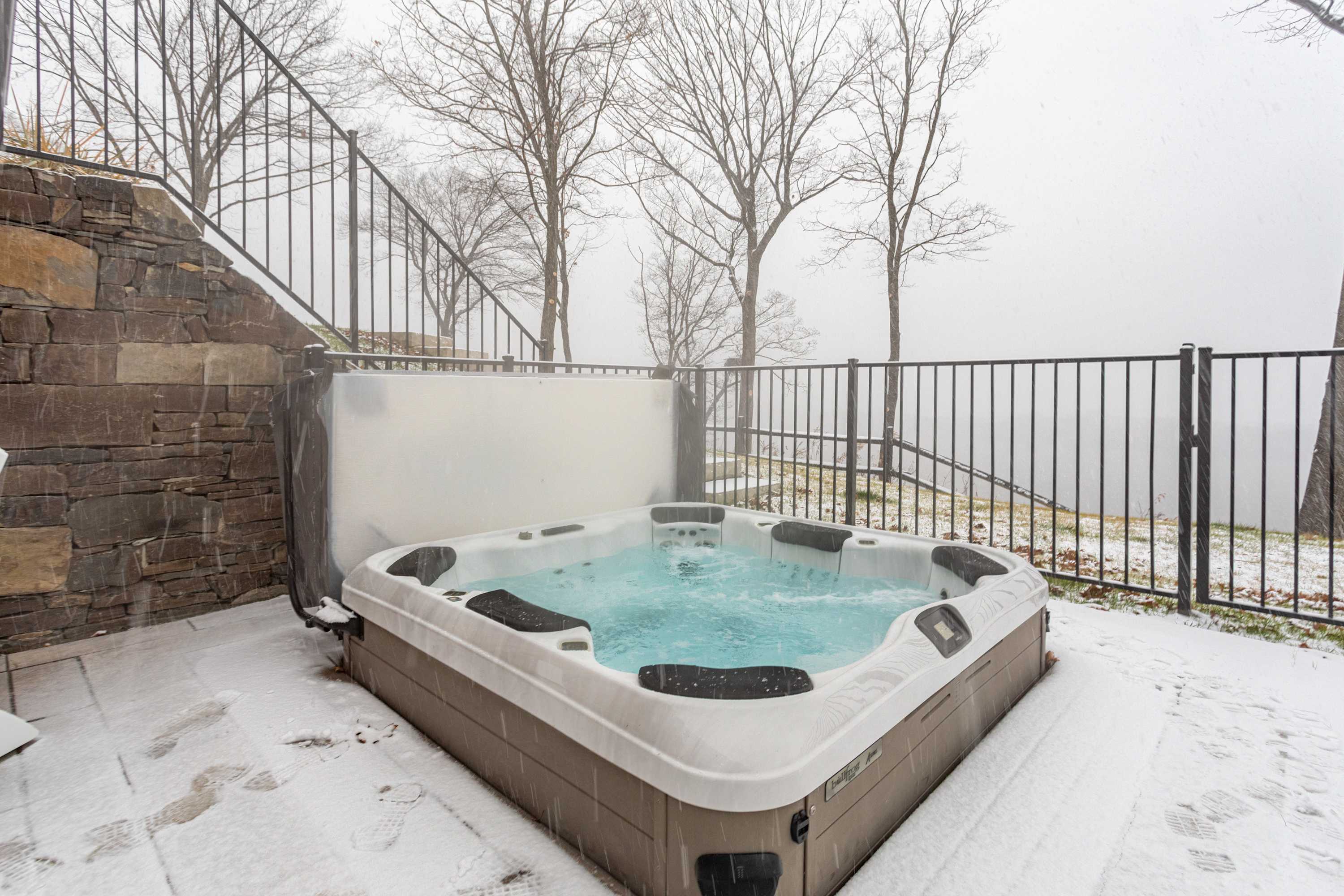 An outdoor hot tub is situated on a snow-covered patio, with a stone retaining wall on the left and a black metal fence enclosing the area. Bare trees and a misty, snowy landscape are visible in the background, creating a serene winter scene.