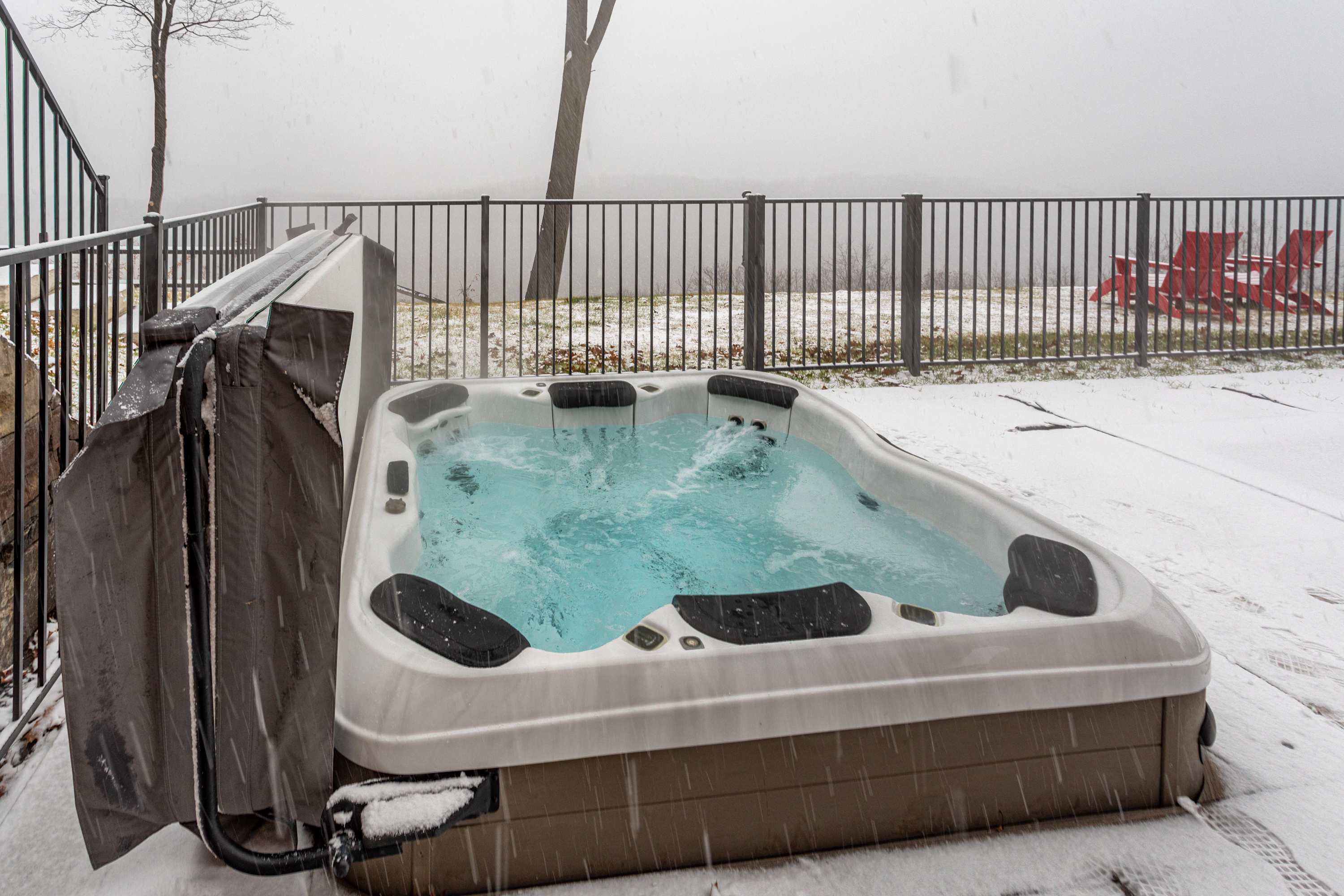A hot tub with its cover partially open and water bubbling is positioned on a snow-covered outdoor patio. A black metal fence runs along the edge of the property, with red Adirondack chairs and a misty, snowy landscape visible in the distance.