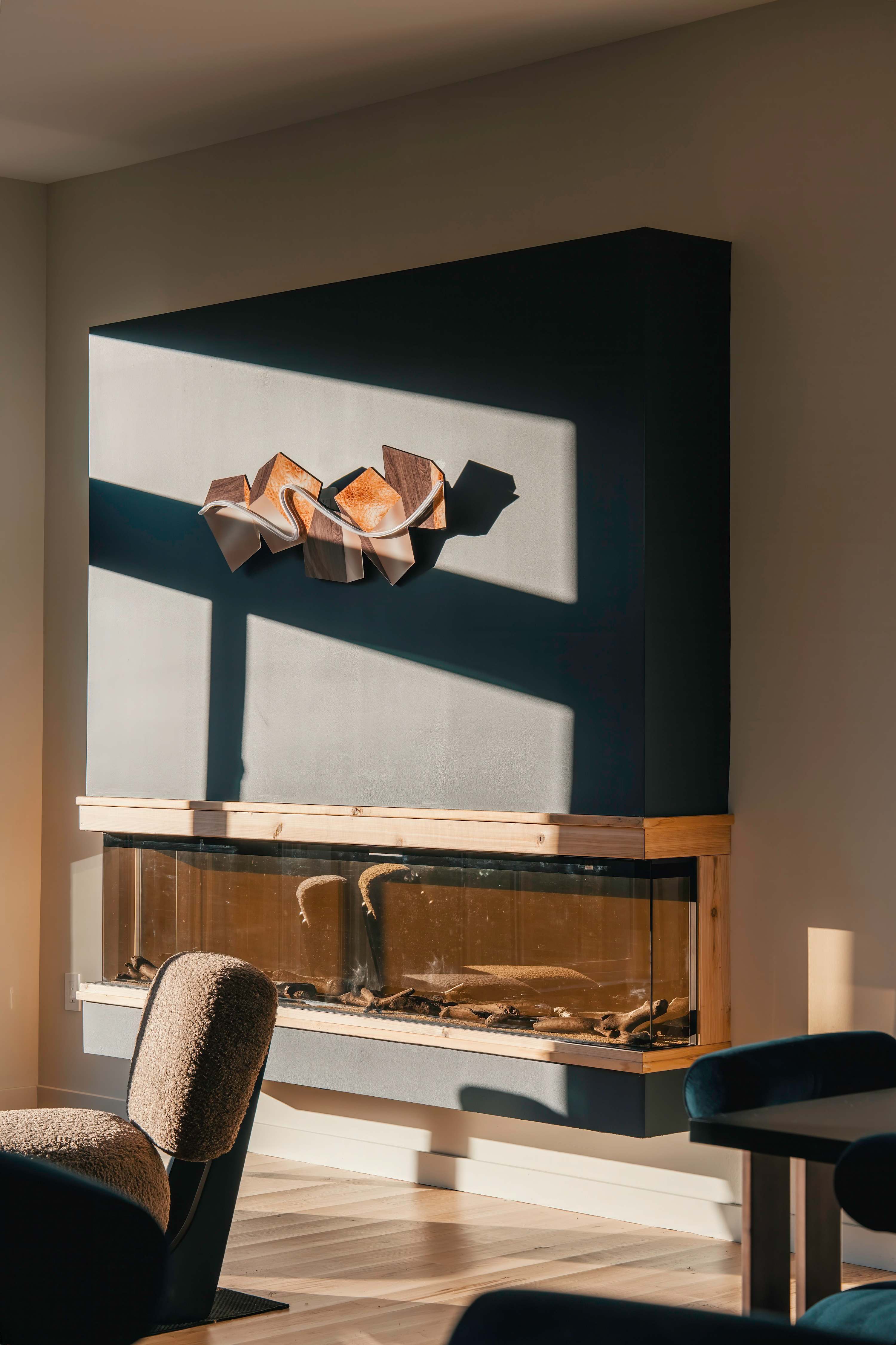 A contemporary living space featuring a dark teal accent wall with a built-in fireplace/aquarium unit, adorned with a sculptural wooden art piece, and illuminated by natural light casting geometric shadows.