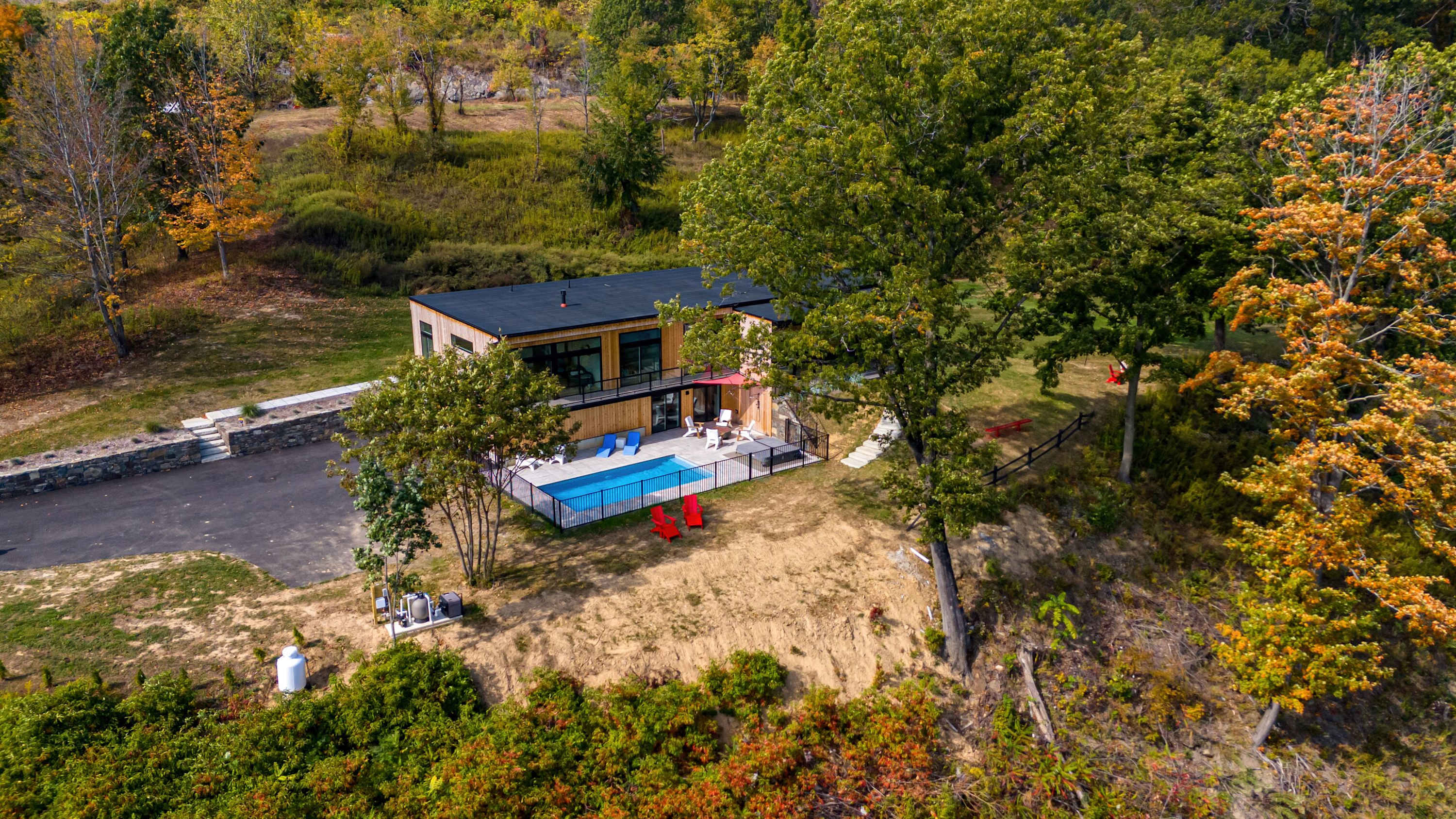 Overhead aerial view of the modern house with a dark roof and light wood siding, situated on a sloped, tree-lined property. The fenced pool area with lounge chairs is visible, along with a paved driveway and surrounding autumnal foliage.