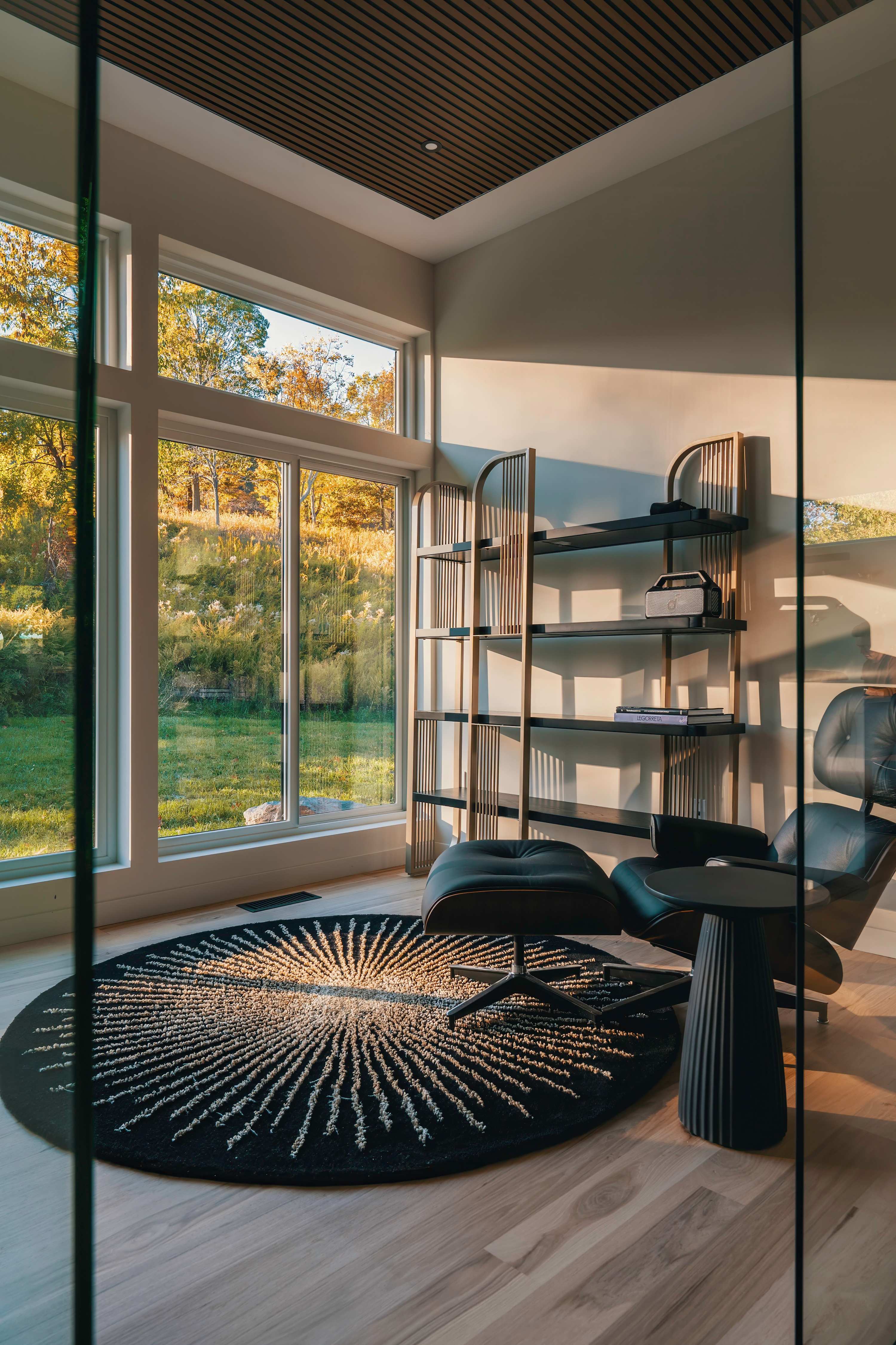 Cozy office nook with a black leather lounge chair and ottoman, a round patterned rug, and a tall gold and black bookshelf, all bathed in sunlight from large windows overlooking a grassy landscape.