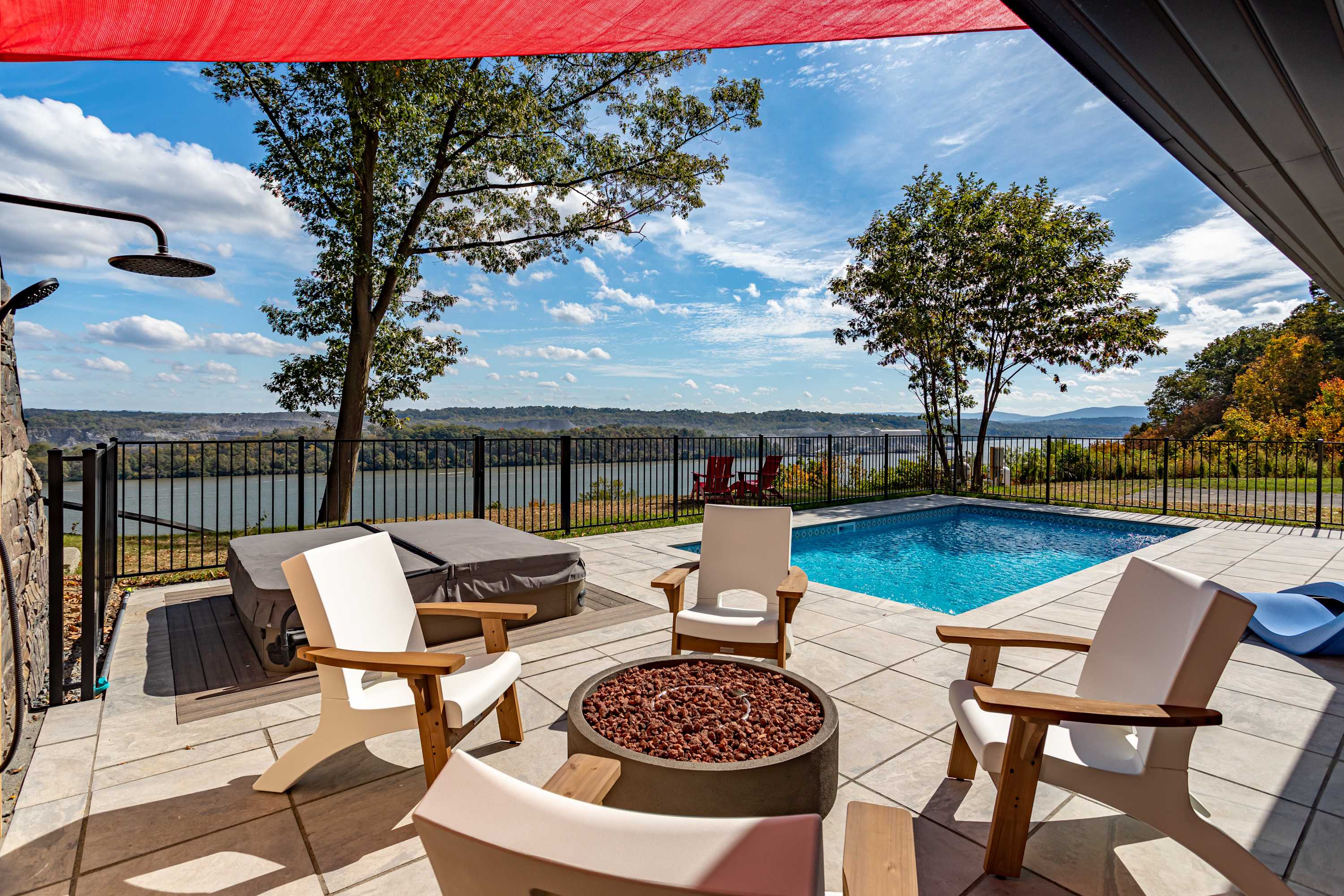 Spacious outdoor patio with a circular fire pit surrounded by modern chairs, a covered hot tub, and an outdoor shower, offering panoramic views of the river and distant landscape under a red shade sail.