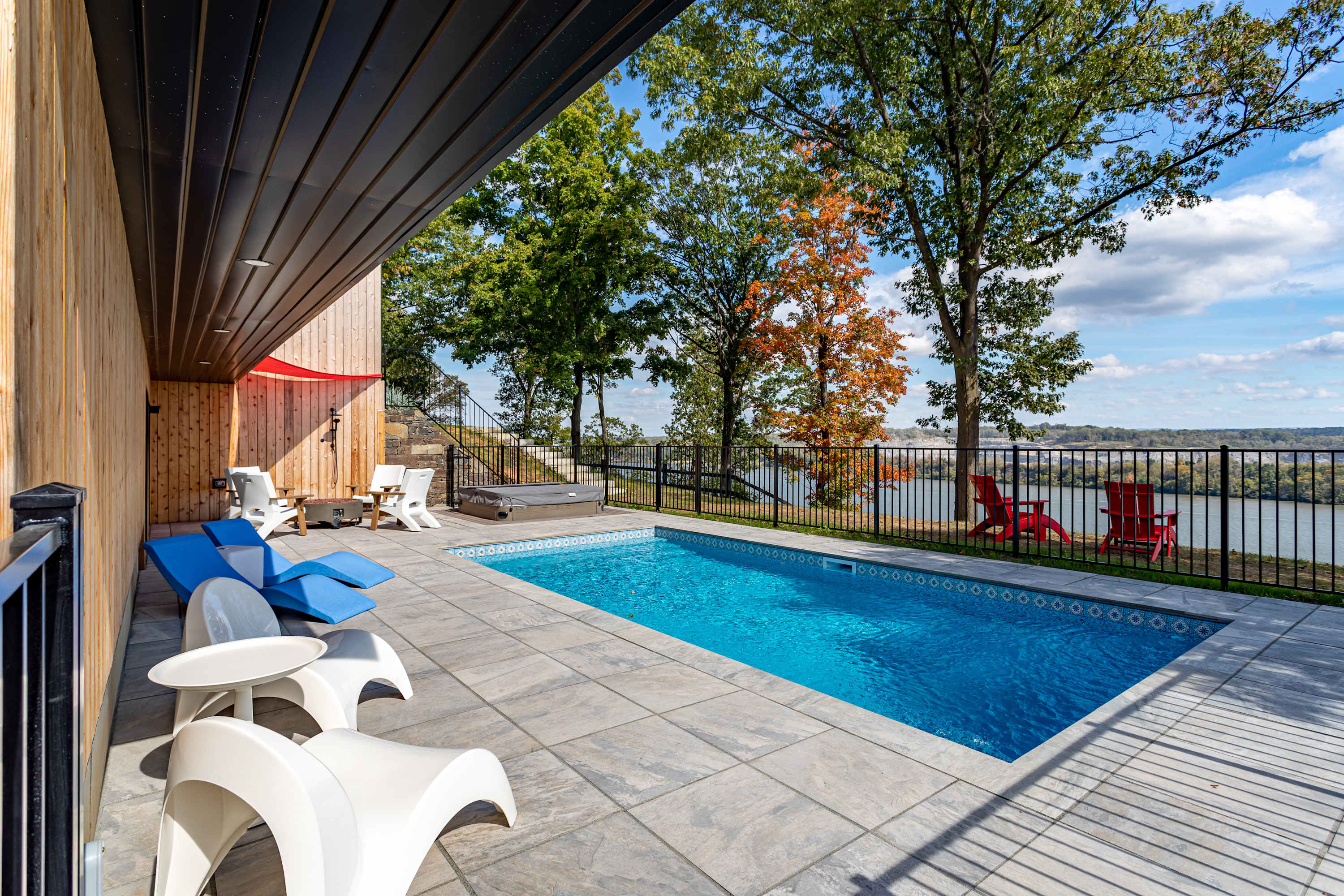 Inviting outdoor pool area featuring a rectangular blue swimming pool, a hot tub, and various lounge seating on light-colored pavers, all set against a backdrop of a modern house, lush trees, and a scenic river view.