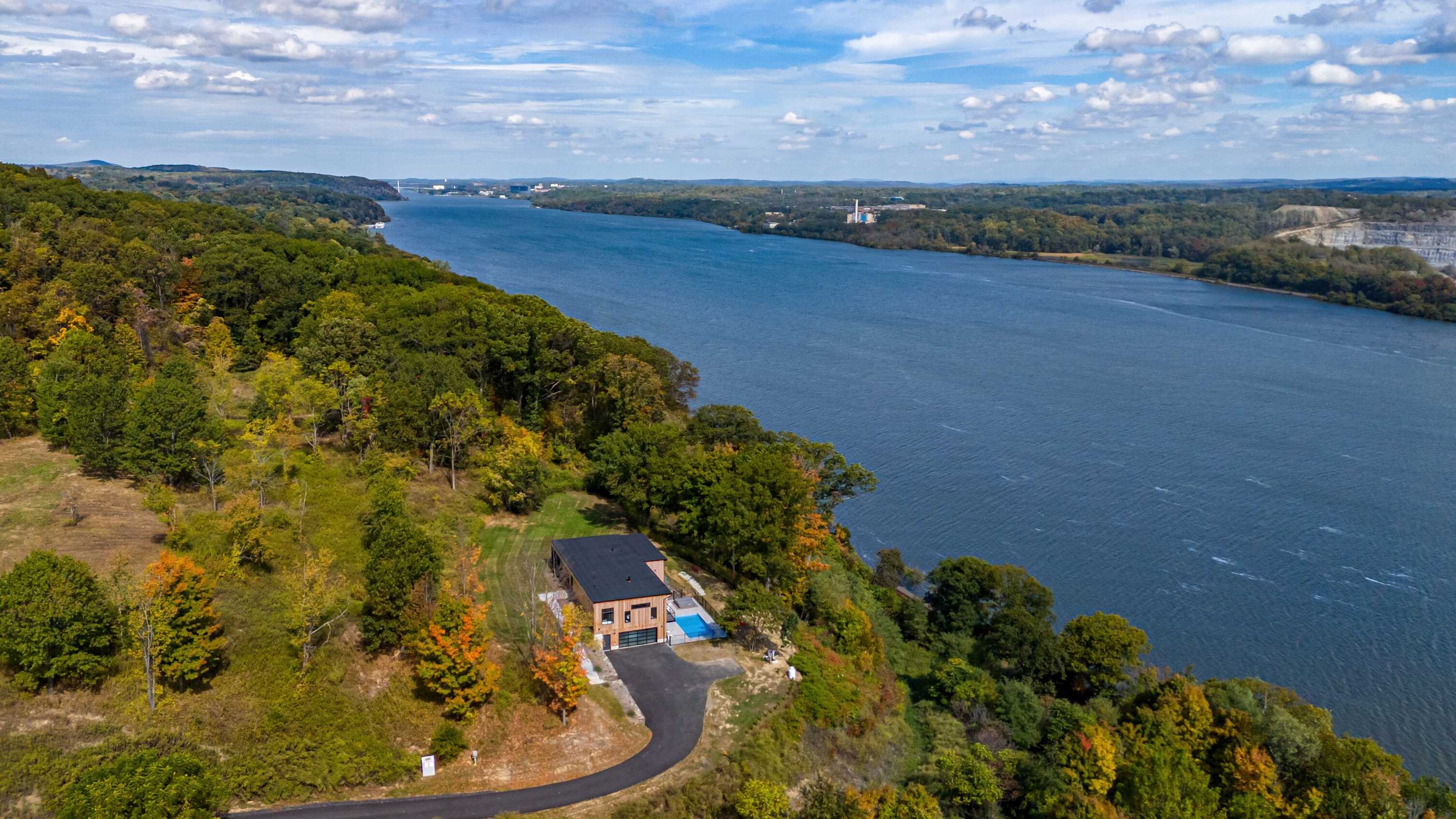 High aerial view showcasing the modern house with a dark roof and light wood siding, nestled on a hillside overlooking a wide river. The property includes a pool area and a winding driveway, surrounded by dense green and autumnal trees.