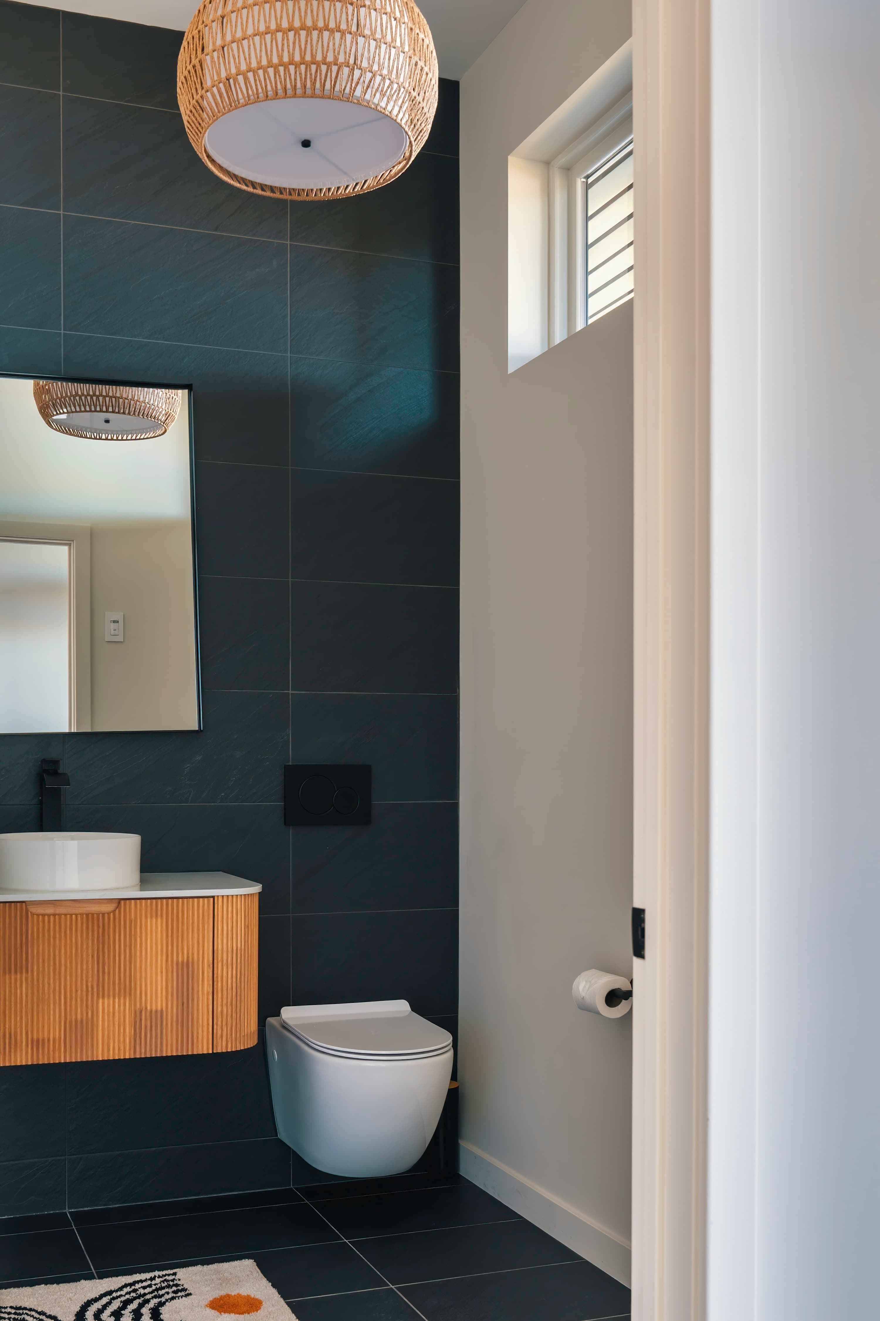 A modern bathroom featuring dark gray tiled walls and floor, a floating wooden vanity with a white vessel sink, a wall-mounted toilet, and a woven pendant light fixture, with natural light from a small window.
