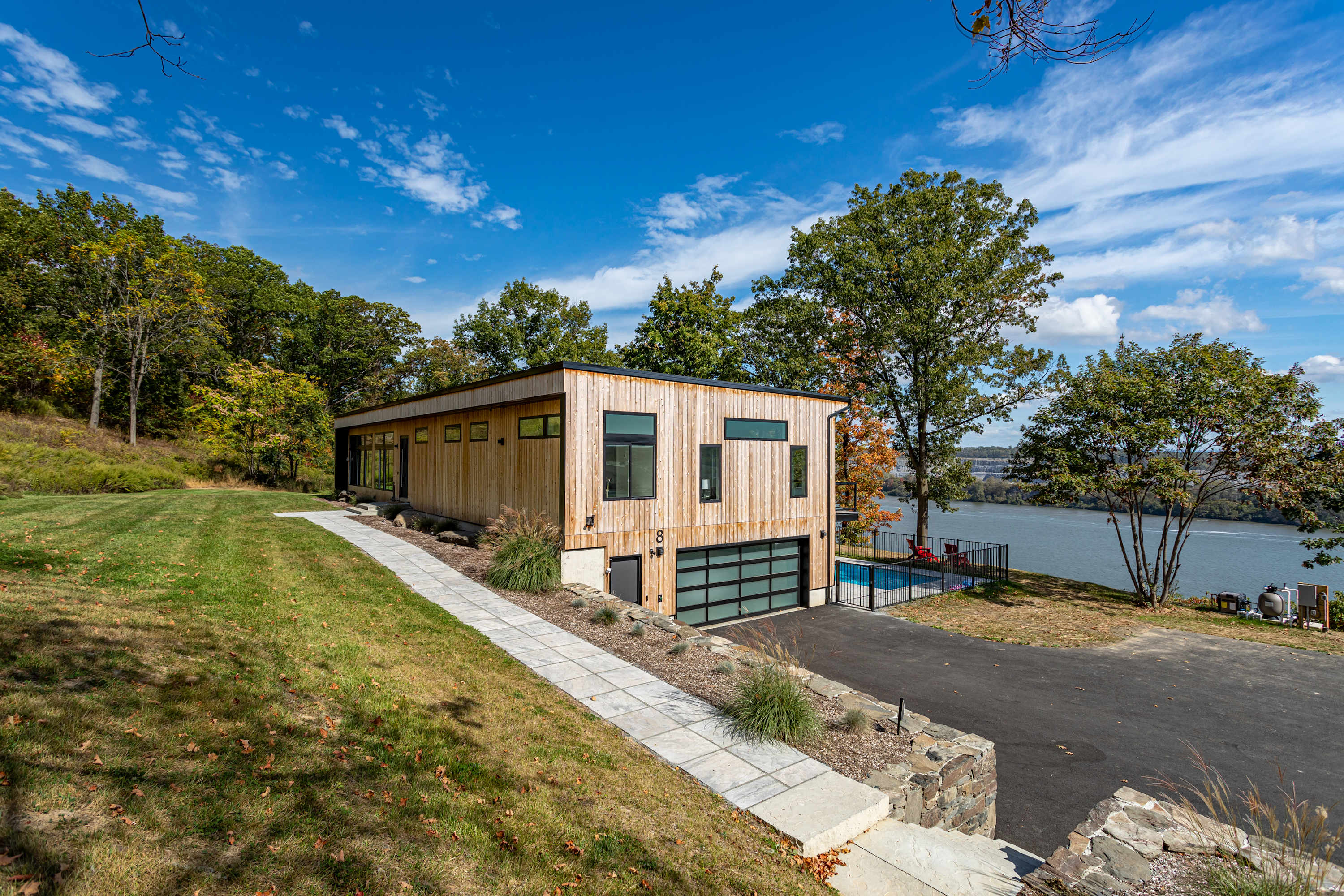 A modern, two-story house with light wood siding and large black-framed windows is nestled on a grassy hillside under a clear blue sky. A paved driveway leads to a garage with a glass door, and a river is visible in the background, along with a small pool area.