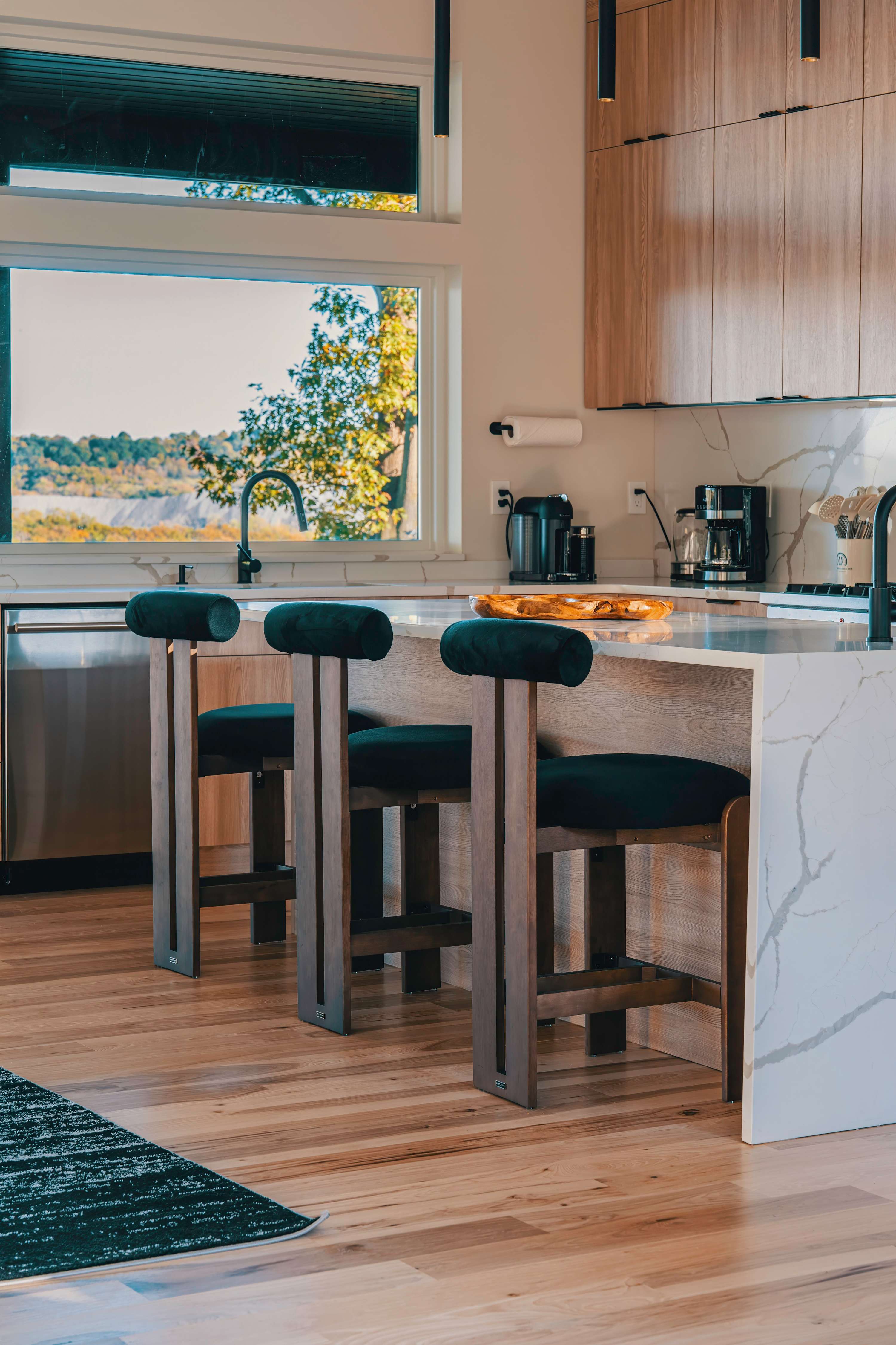 A modern kitchen island with a white quartz countertop and waterfall edge, featuring three dark green velvet bar stools with unique wooden bases, and a view of a lush green landscape through a large window.