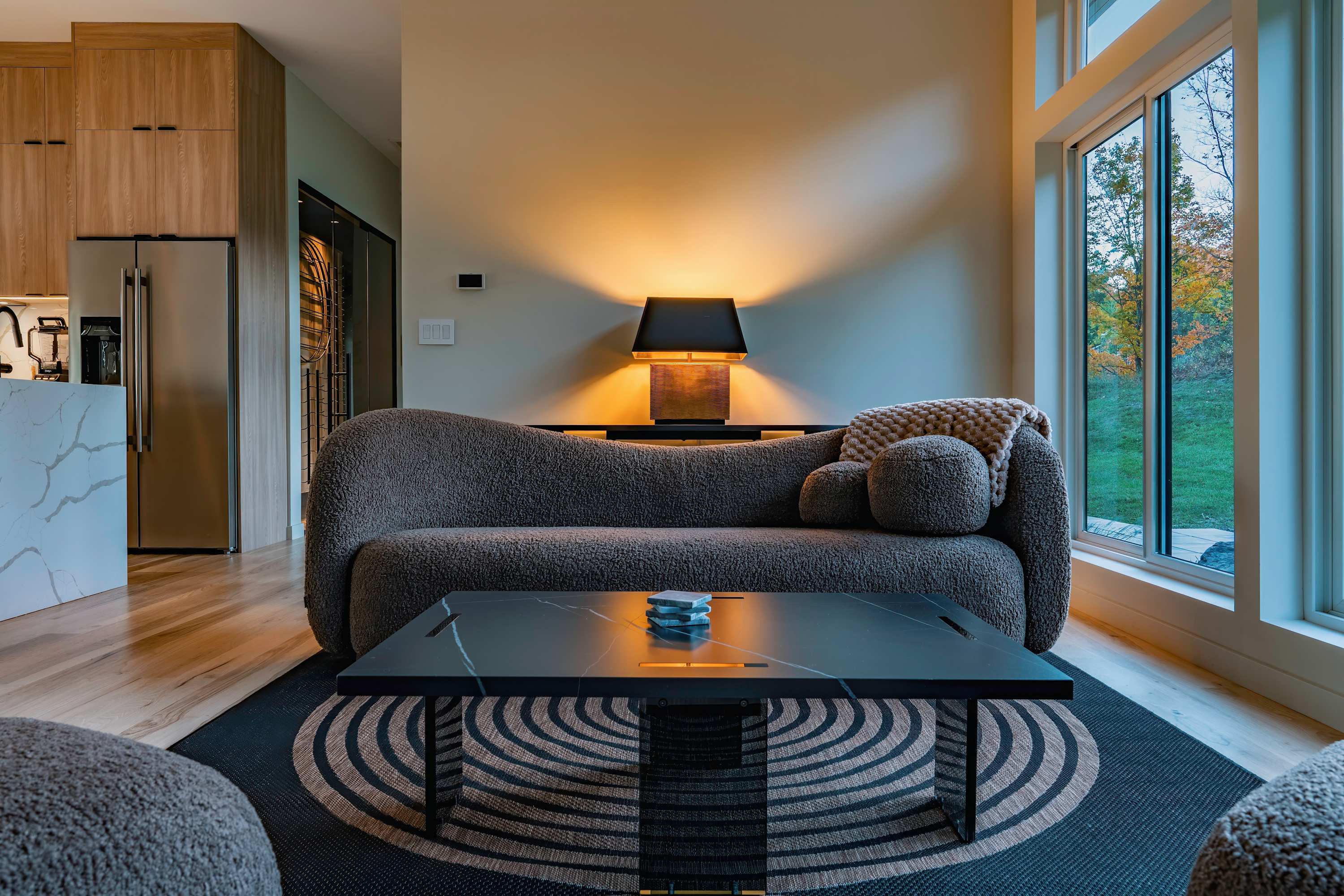 A cozy living area featuring a curved, textured brown sofa with a matching throw and pillows, a black marble-top coffee table, and a patterned rug, all bathed in the warm glow of a table lamp.