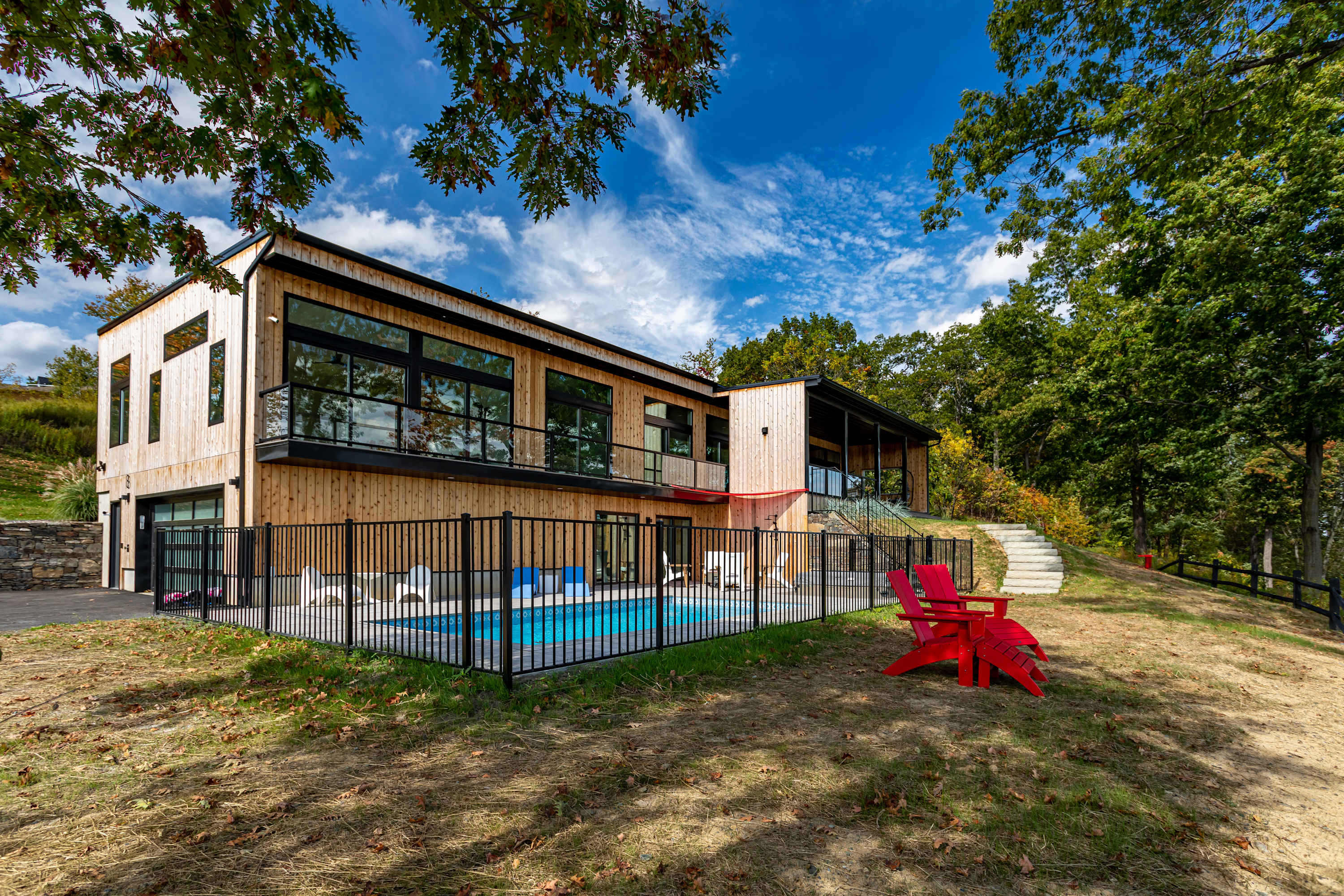 Side exterior view of the modern wood-sided house with large windows, overlooking a fenced pool area with white lounge chairs and red Adirondack chairs on the grassy lawn. A stone staircase leads up the hill in the background.