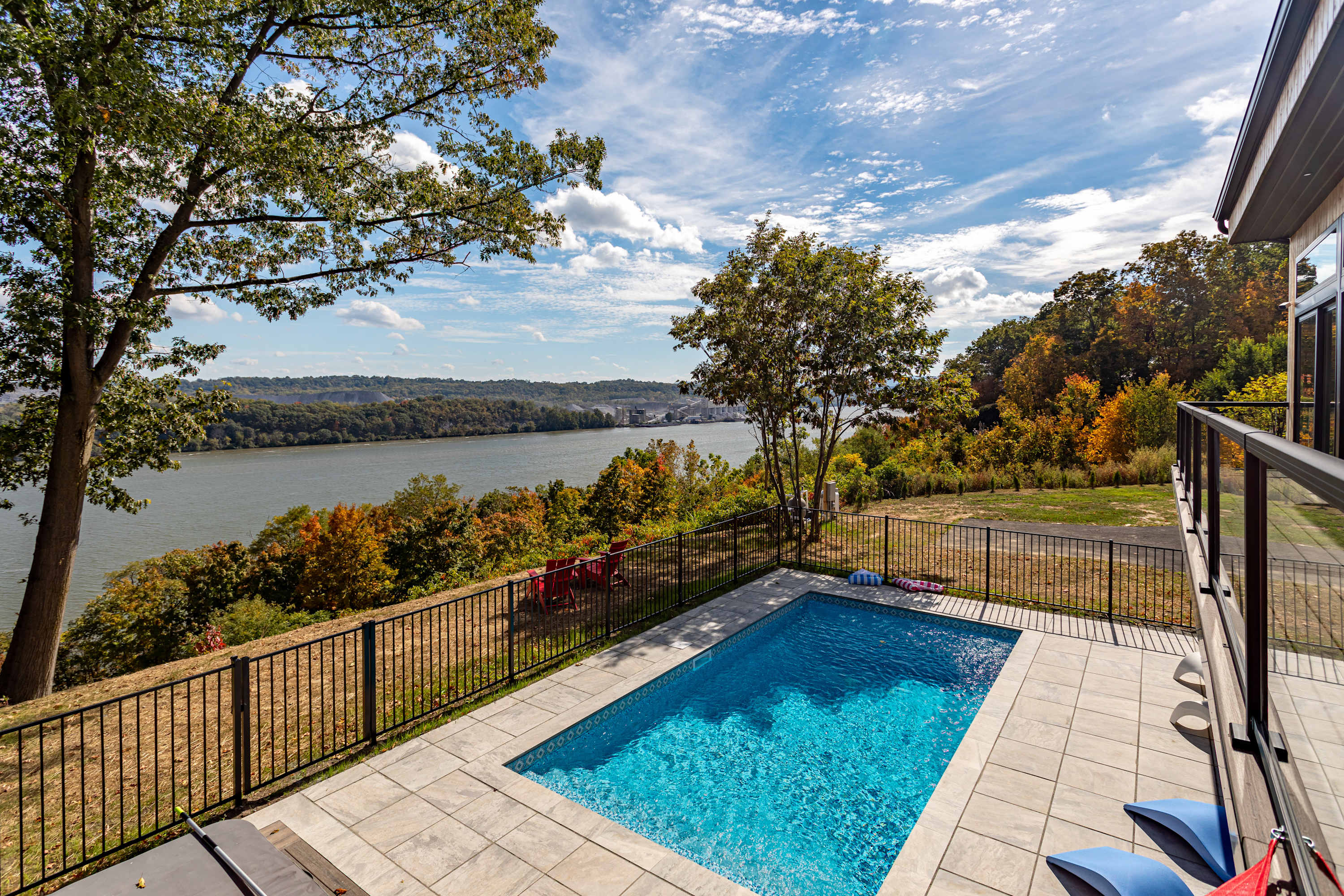 Rectangular outdoor swimming pool with blue water, surrounded by light-colored paving stones and a black metal fence. The pool area offers stunning panoramic views of a wide river and lush, colorful trees.