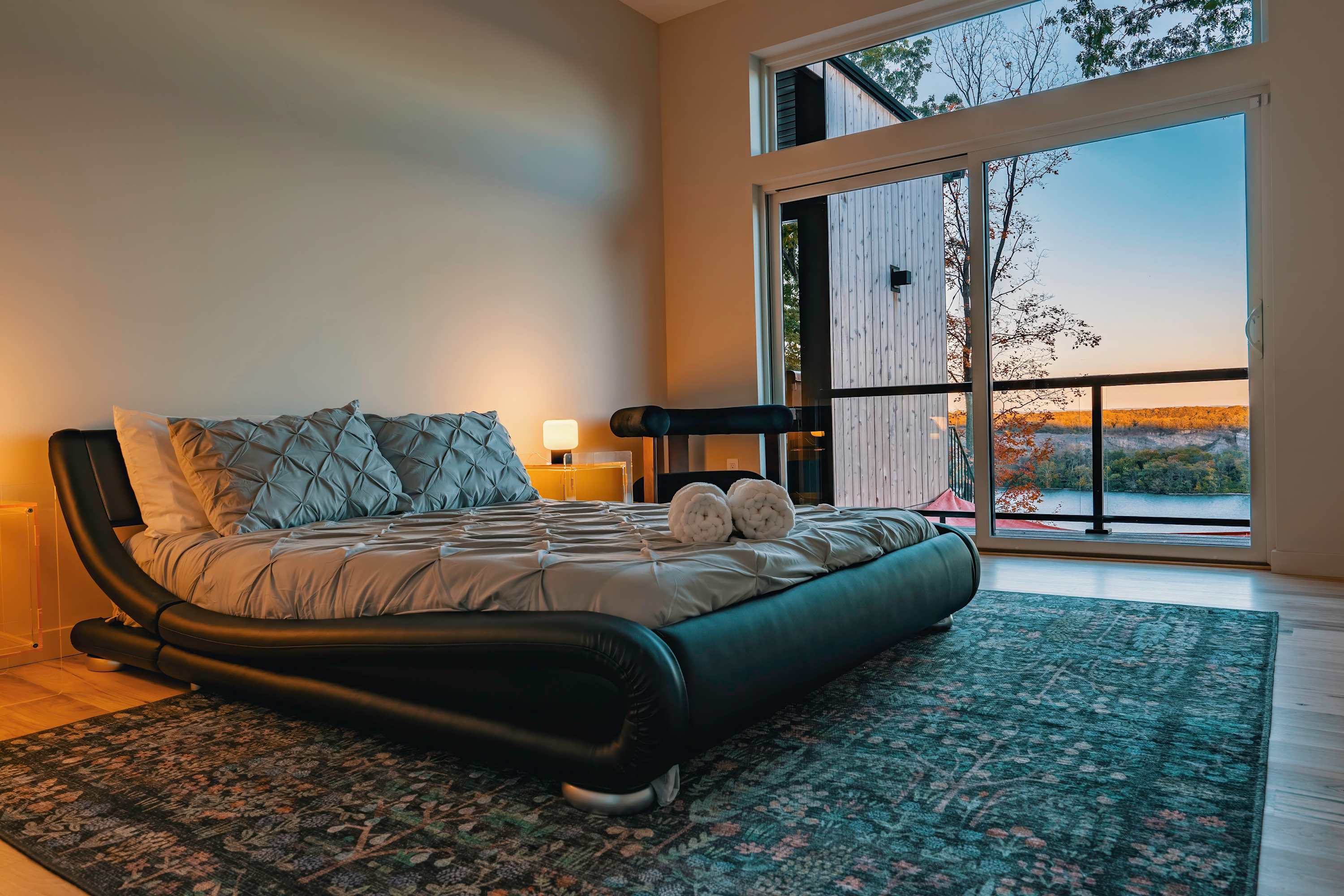 Stylish bedroom with a unique black curved platform bed, a patterned area rug, and floor-to-ceiling windows offering stunning views of a lake and autumn foliage.