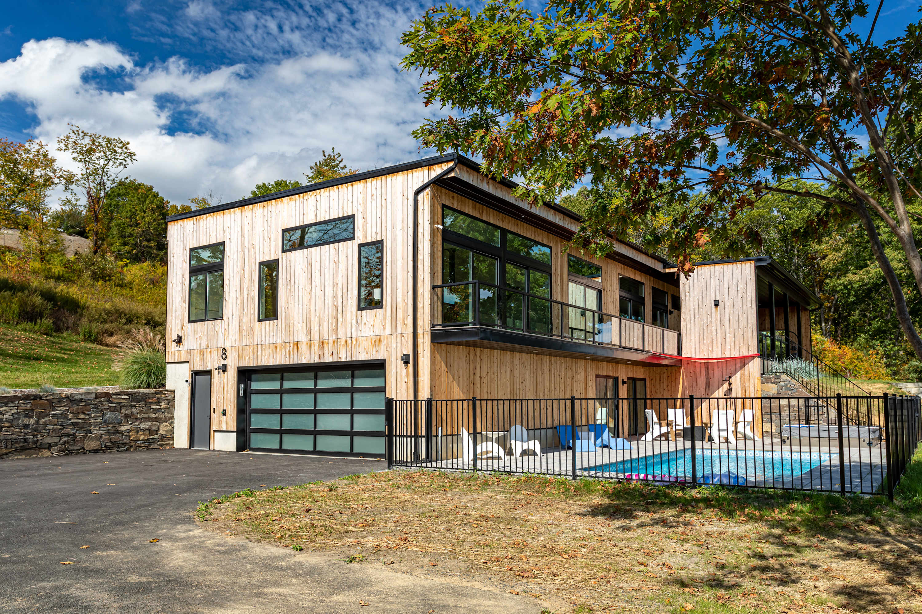 Modern two-story house with light wood siding and large black-framed windows, featuring a glass garage door and a fenced pool area to the right. A paved driveway leads to the entrance, surrounded by trees and a stone wall.