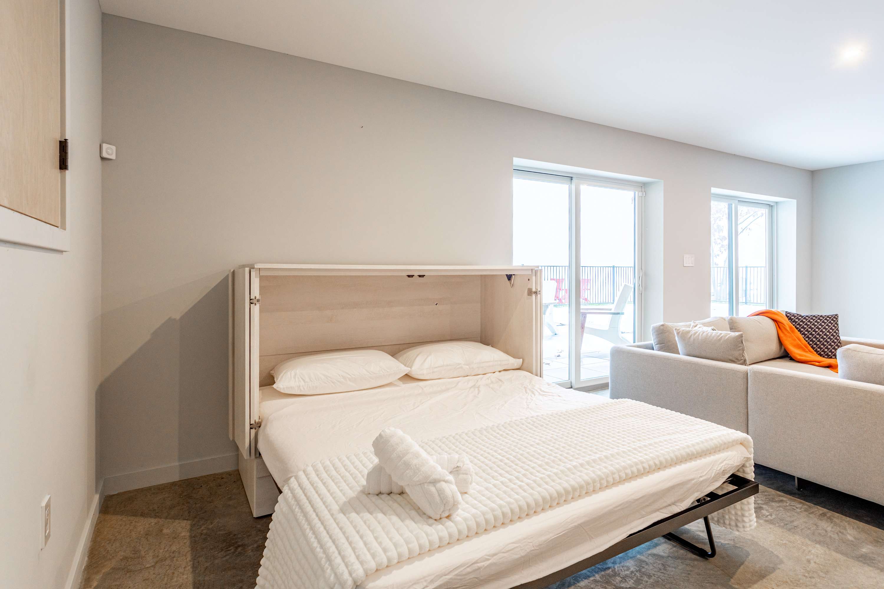 A close-up view of a white fold-out bed with crisp white bedding and rolled white towels, positioned against a light gray wall. The bed frame is integrated into a white cabinet, and a sliding glass door is visible in the background.