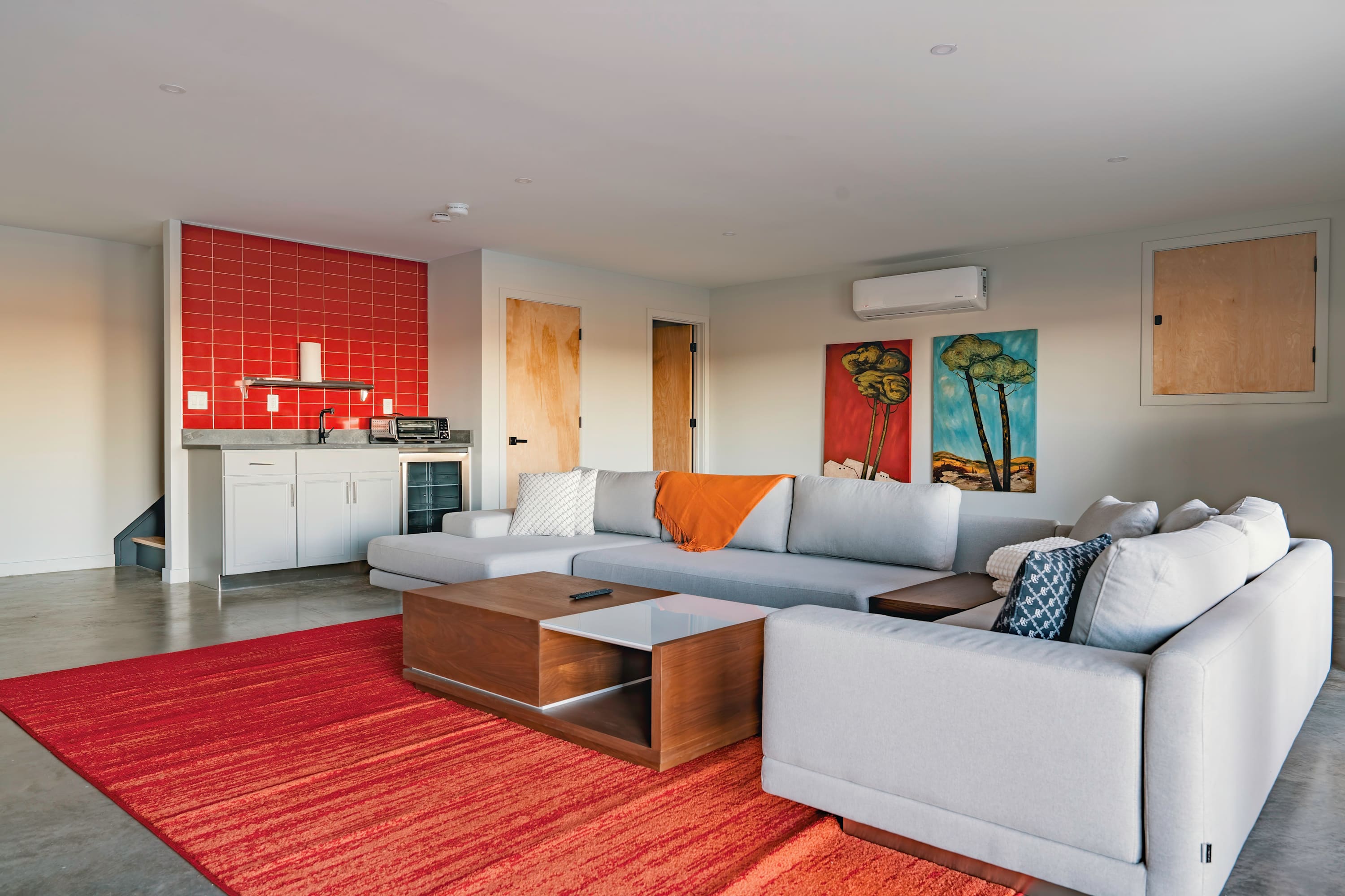 A vibrant secondary living space or game room featuring a large light gray sectional sofa, a wooden coffee table, a bright red area rug, and a compact kitchenette with red tiled backsplash, offering a lively and functional area.