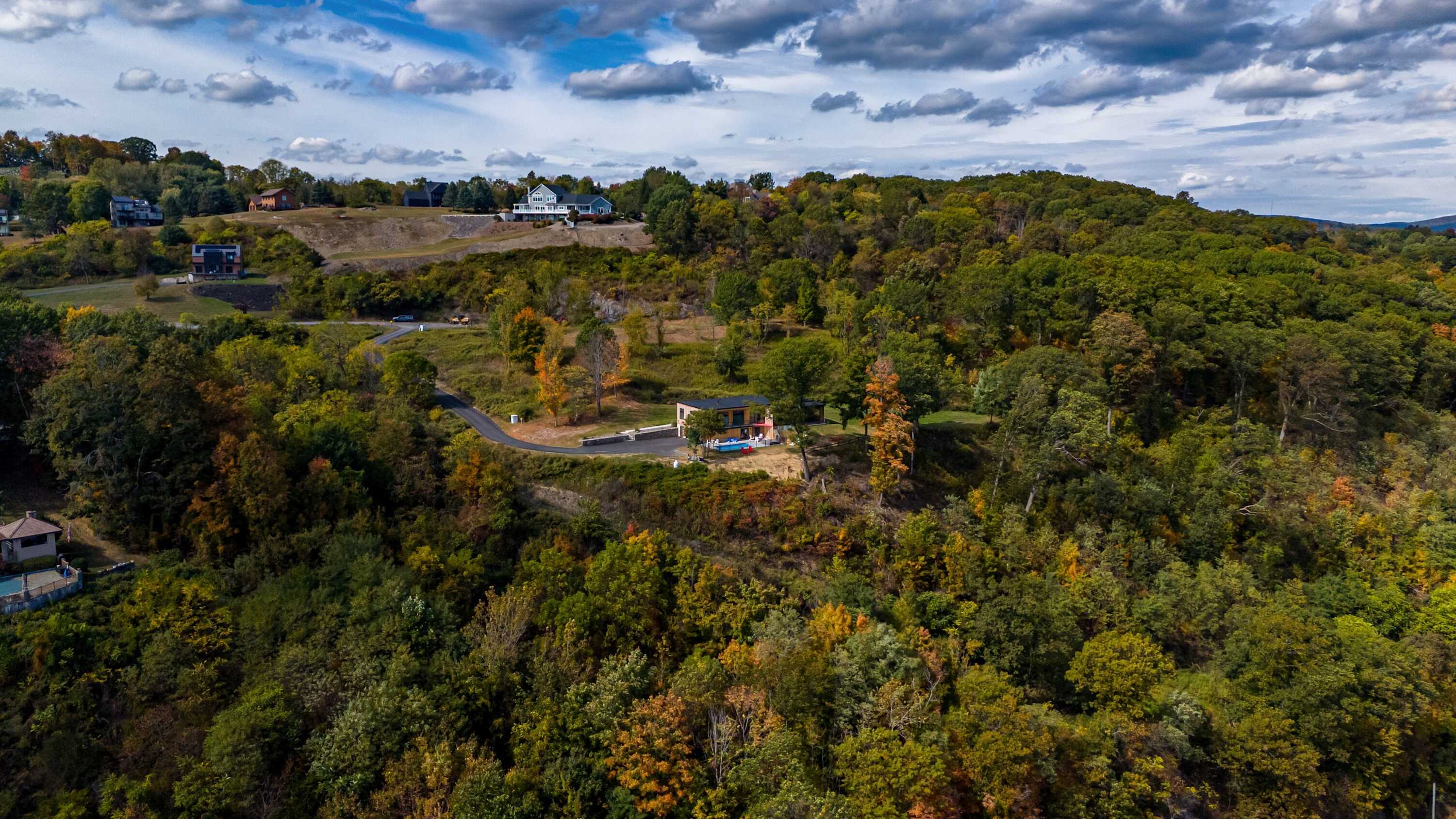 Expansive aerial view of the modern house nestled on a lush, tree-covered hillside, with a winding road leading up to it. The property offers breathtaking panoramic views of a wide river and distant hills under a partly cloudy sky.