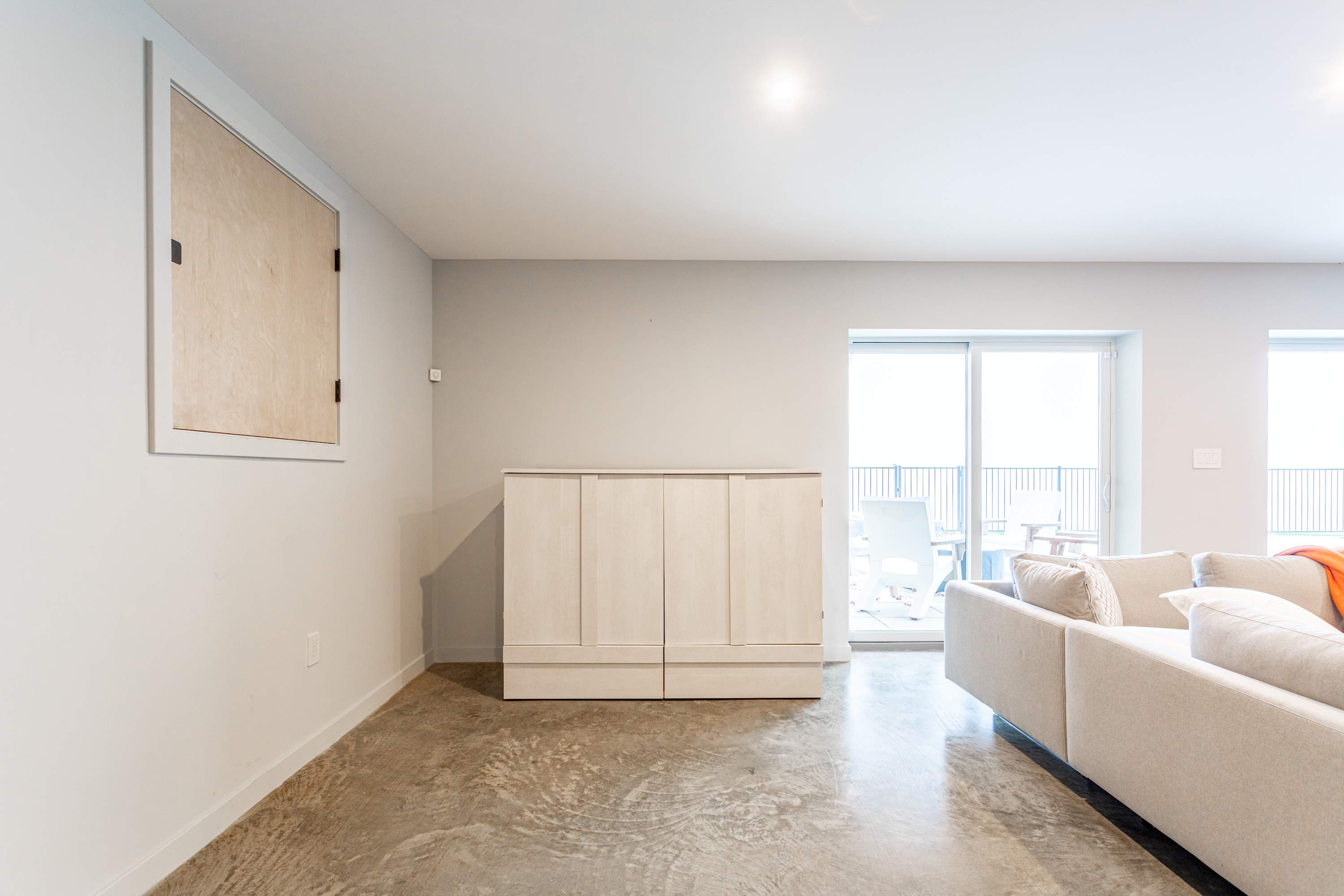 A minimalist corner of a secondary living space with light gray walls and a concrete floor, featuring a closed white cabinet that conceals a murphy bed, next to a sliding glass door leading to an outdoor balcony.
