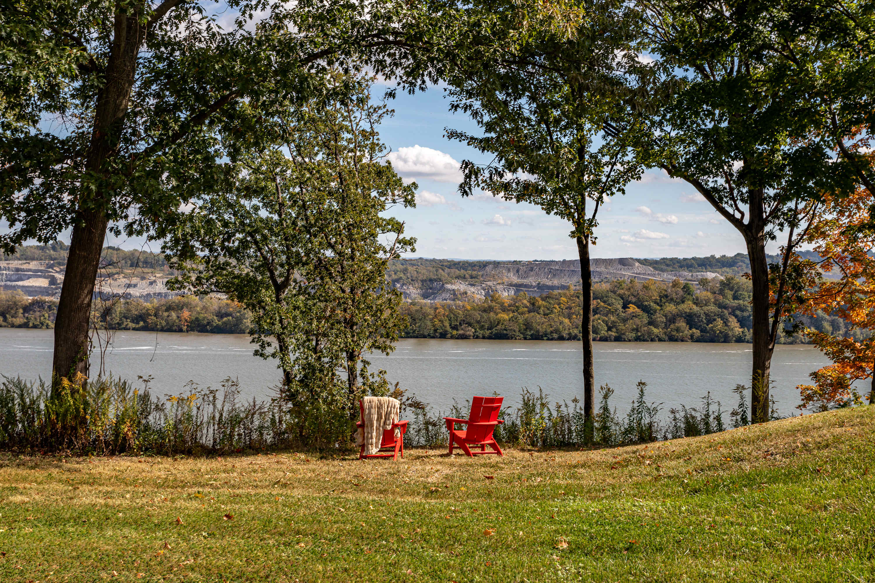 Scenic outdoor spot with two red Adirondack chairs placed on a grassy knoll, framed by trees, offering a serene view of the wide river and the distant quarry on the opposite bank.