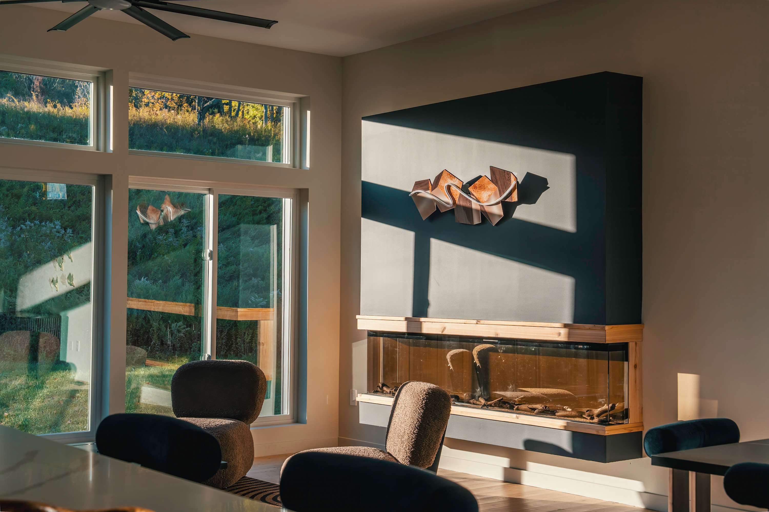 Modern living space featuring a dark accent wall with a unique sculptural art piece above a linear gas fireplace with a glass-enclosed aquarium-like base, complemented by large windows offering views of lush greenery.