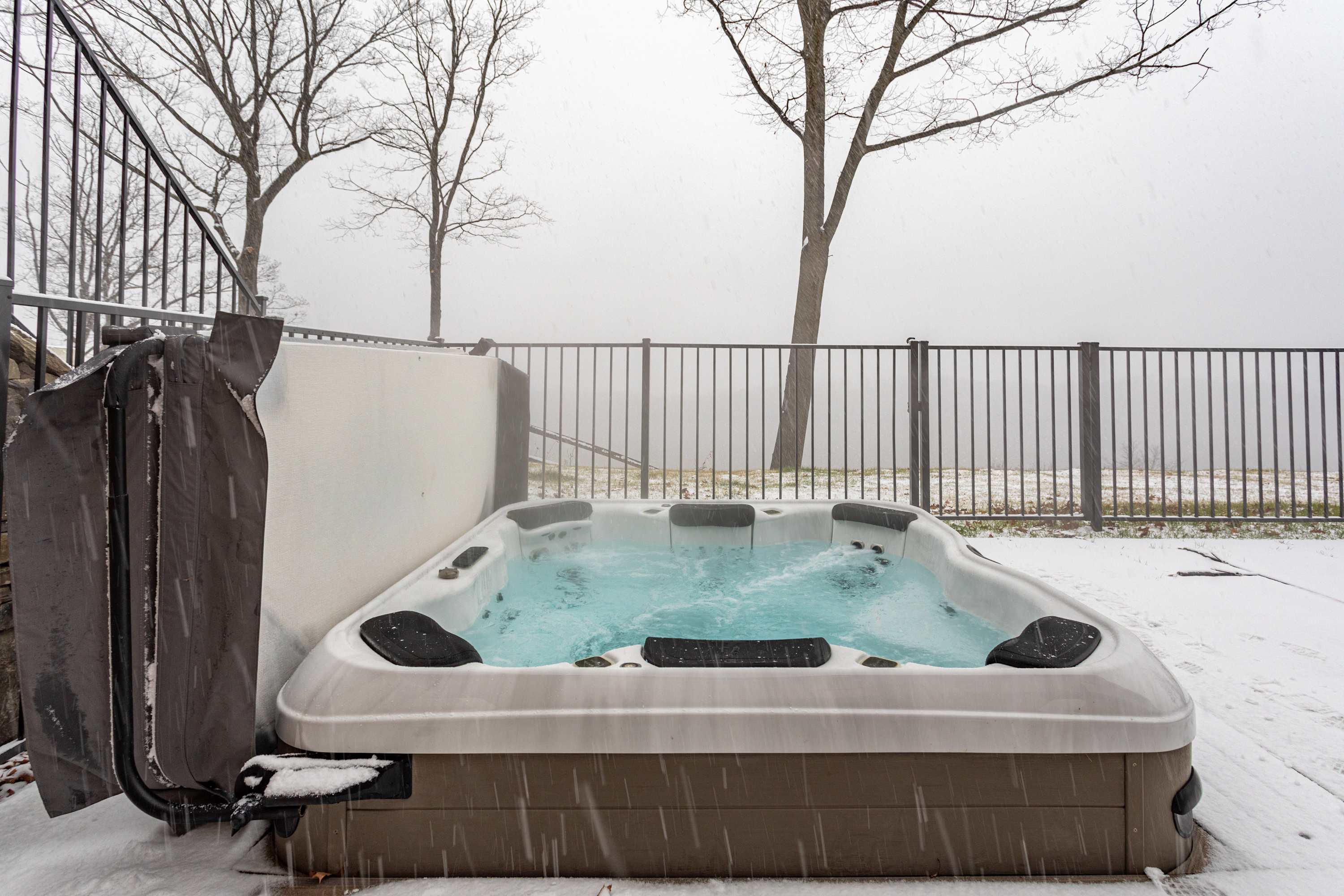 A hot tub with active jets is centered on a snow-covered outdoor patio, framed by a black metal fence and bare trees. The misty winter weather creates a tranquil backdrop, emphasizing the warmth of the bubbling water.