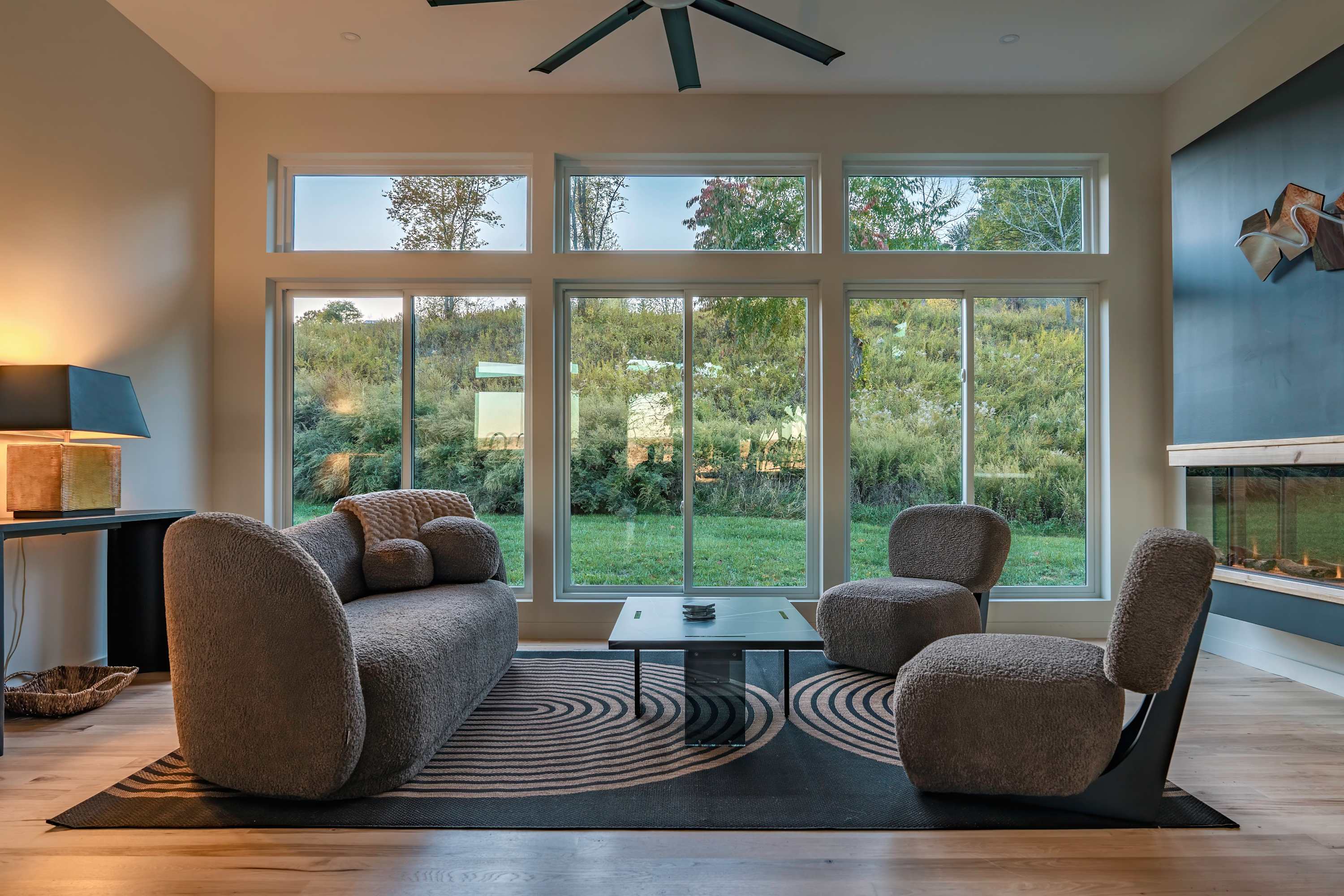 Cozy living room featuring a modern gray sofa and two armchairs with textured upholstery, a black coffee table, and a patterned rug, set against a wall with a fireplace and large windows offering a view of a grassy hillside.