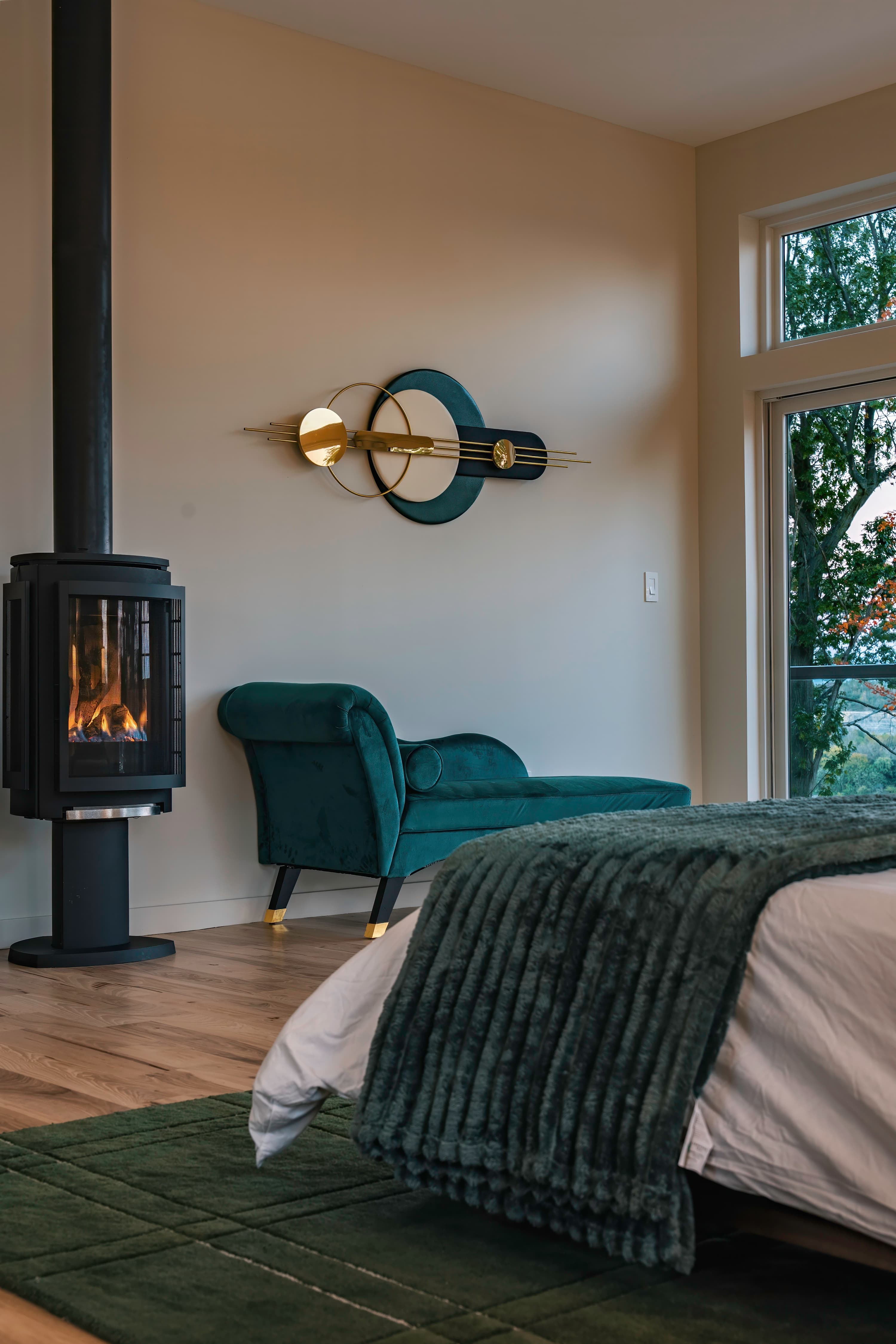 Cozy corner of a bedroom featuring a modern black gas fireplace, a teal velvet chaise lounge, and a decorative wall art piece, with a large window providing views of the outdoors.