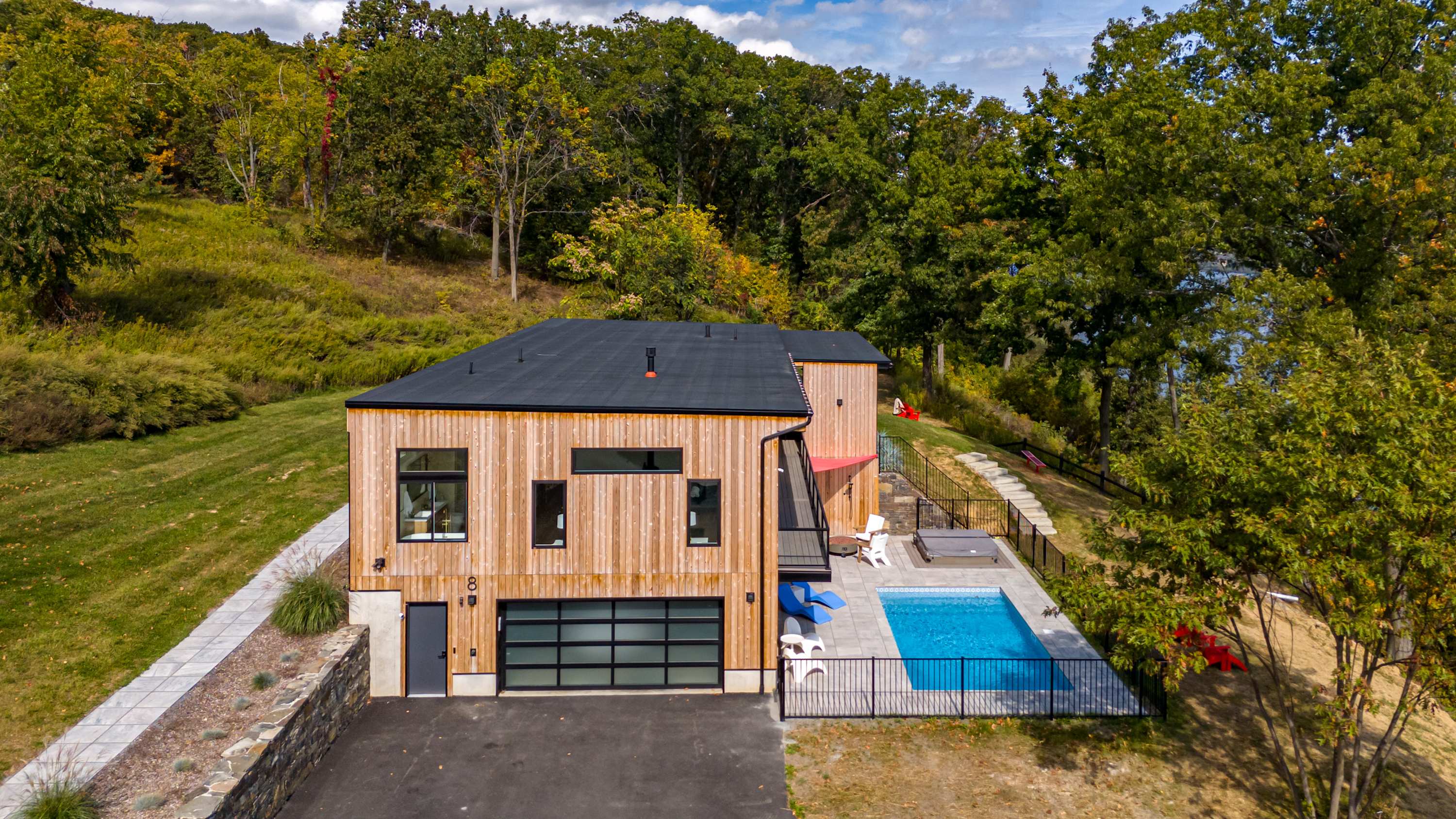 Exterior view of the modern house with light wood siding and a dark roof, featuring a glass garage door and a fenced pool area with a hot tub. The property is surrounded by lush trees and a paved driveway.