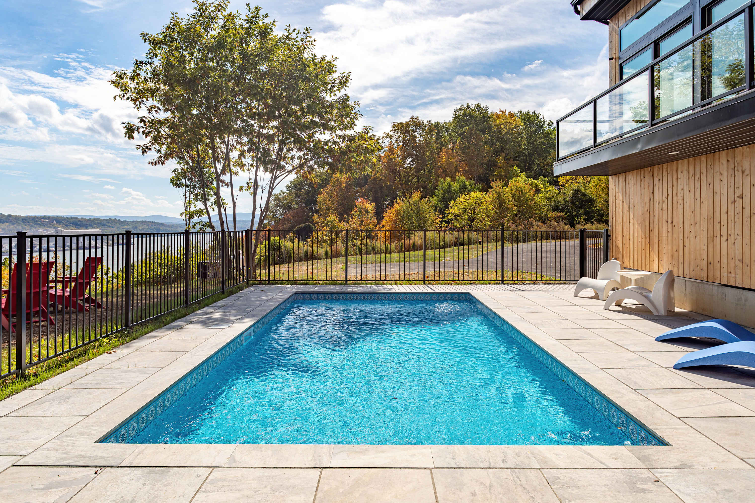 Close-up view of the rectangular outdoor swimming pool with blue water, surrounded by light paving and a black fence. White and blue lounge chairs are placed on the patio, with the modern wood-sided house visible on the right.