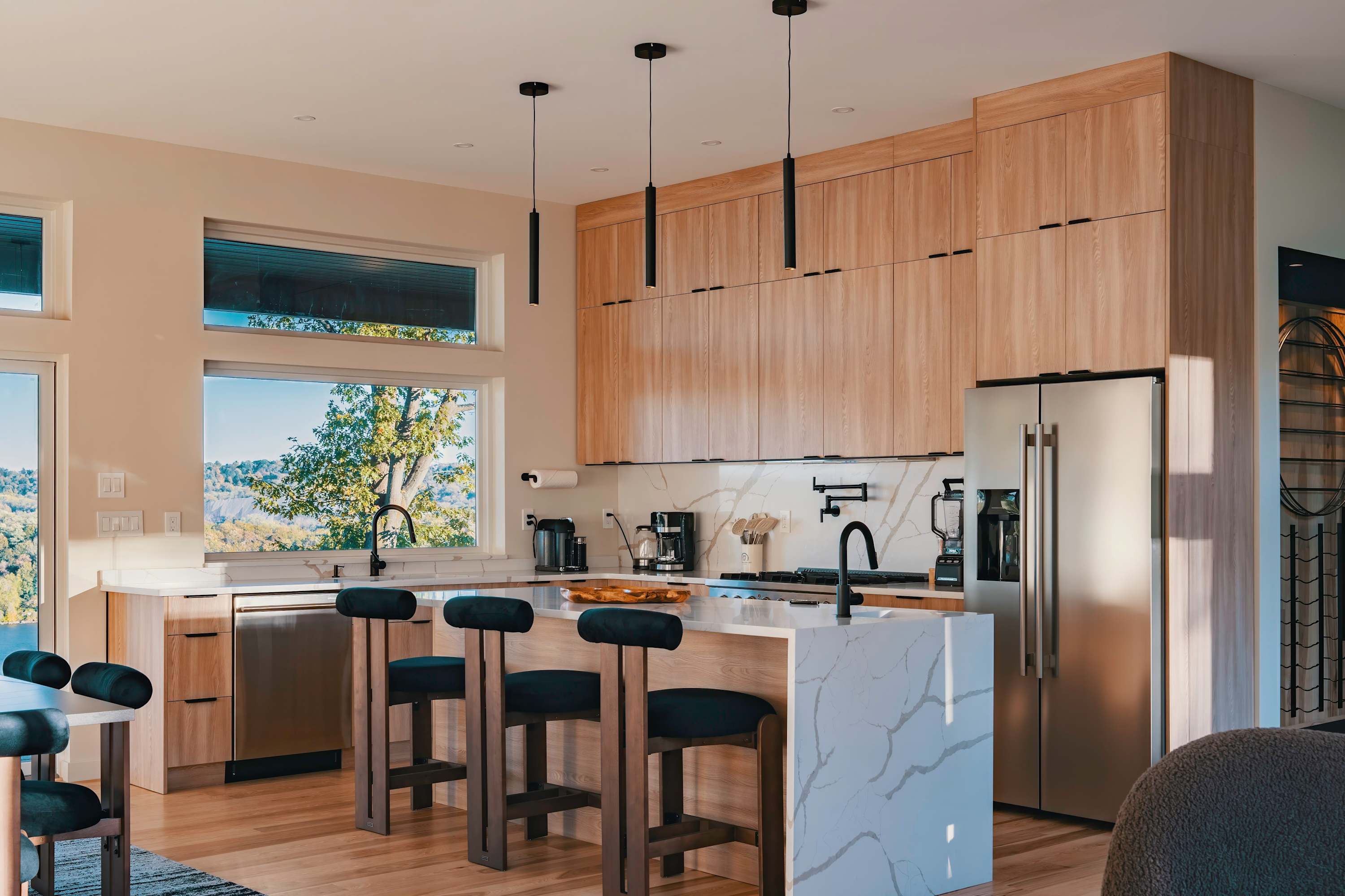A spacious, modern kitchen with light wood cabinetry, white quartz countertops, and a large island with three dark green bar stools, offering panoramic views of a natural landscape through expansive windows.