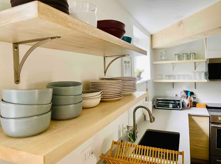 Detailed view of the modern kitchen featuring light wood open shelving stocked with various dishes and bowls. A stainless steel sink with a gooseneck faucet and a wooden dish rack are visible on the light gray countertop.