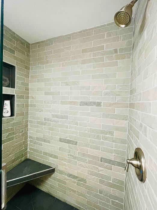 A modern shower stall features light grey brick-patterned tile walls and a built-in bench. A brushed nickel shower head and control knob are visible, along with a recessed niche holding toiletries.