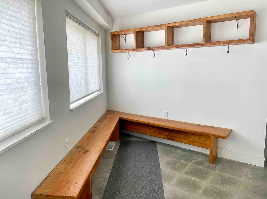 A bright mudroom features a long, L-shaped wooden bench along two walls, with a dark grey rug on the concrete floor. Above the bench, a rustic wooden shelf unit with cubbies and metal hooks is mounted on the white wall, beneath two large windows with blinds.