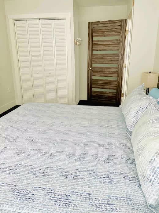View of bedroom-1 showing a white louvered bifold closet door and a modern wooden door with horizontal glass panels. The light blue and white striped bedding of the queen bed is visible in the foreground.