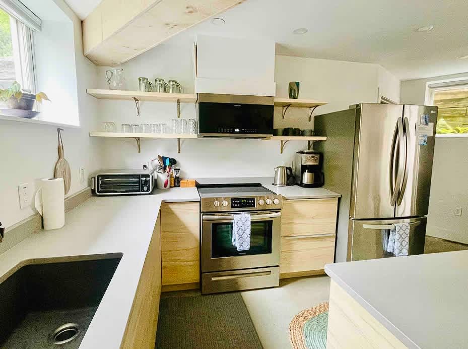 Modern kitchen equipped with stainless steel appliances including a stove, microwave, and refrigerator, set against light wood cabinetry and light gray countertops. Open shelving above the counter holds glasses and small decor items.