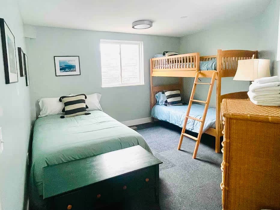 A light green-walled bedroom features a single bed with a dark green duvet and a striped pillow, alongside a wooden bunk bed with light blue bedding. A window with blinds is centered, and a dark wooden chest sits at the foot of the single bed.